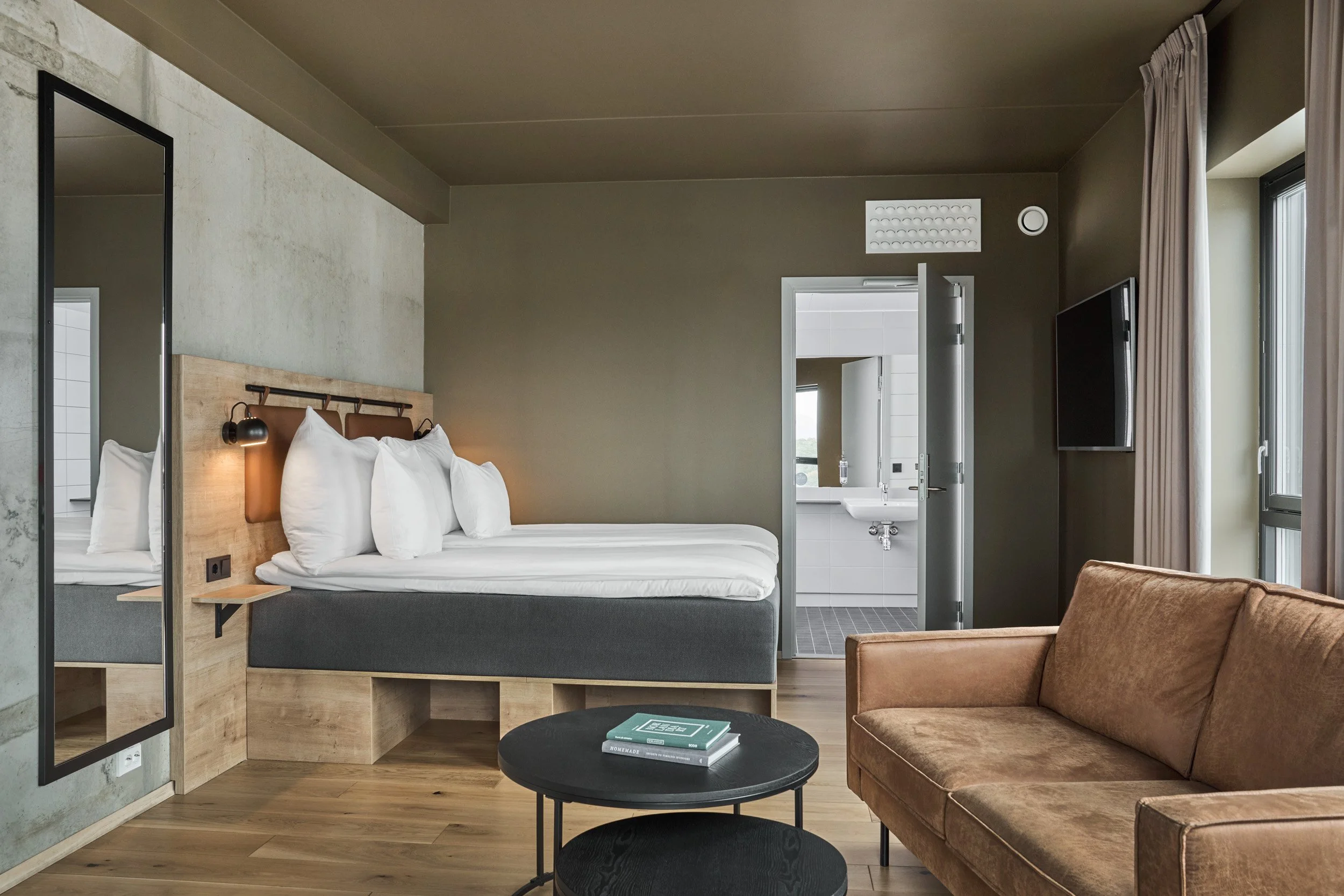 A modern hotel room with a bed, a brown sofa, a round black coffee table, a wall-mounted TV, and a doorway leading to a bathroom with a sink and toilet, large windows with curtains, and wooden flooring.