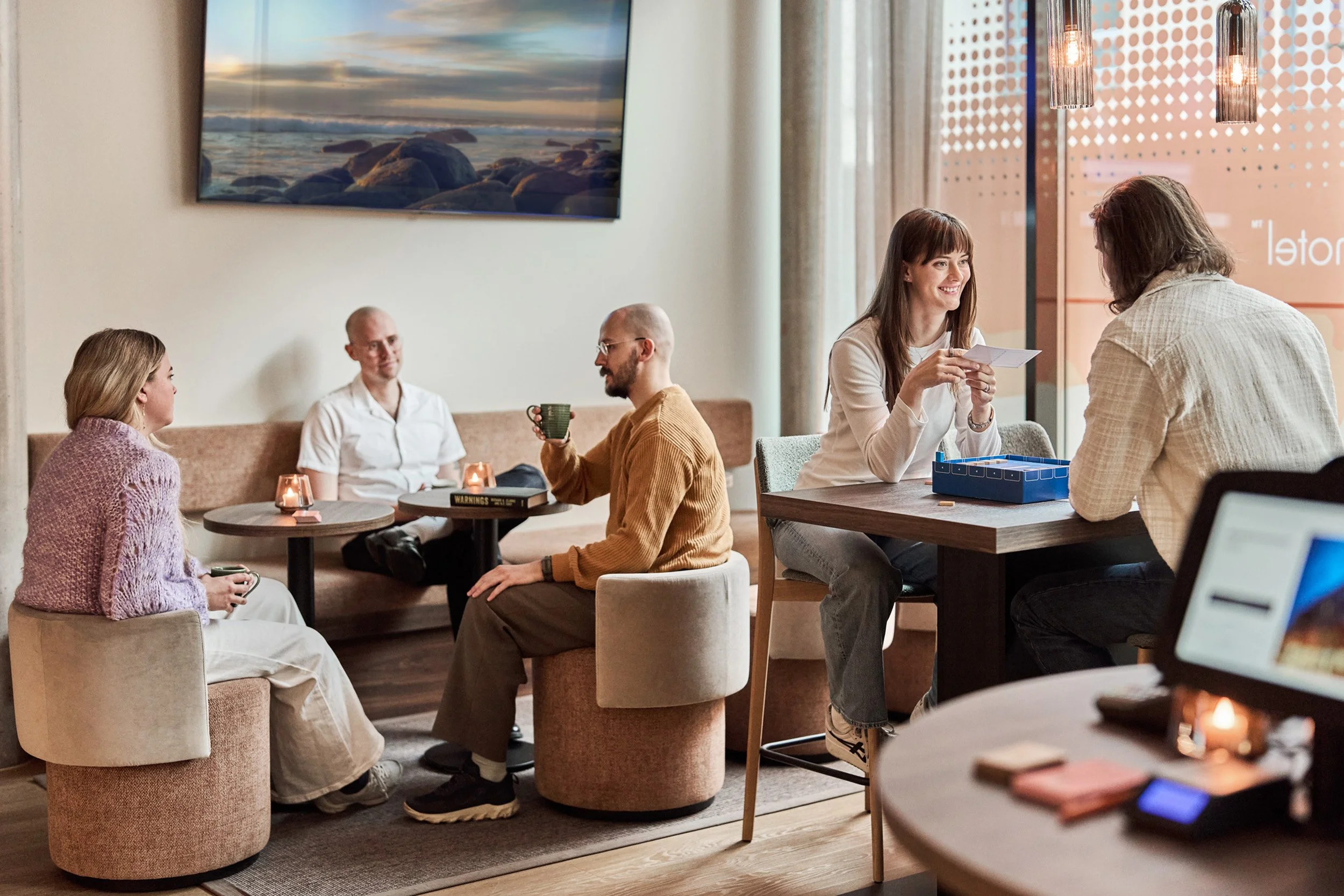 People sitting in a cozy cafe, engaging in conversations and working on laptops, with warm lighting and a large screen showing a nature scene on the wall.