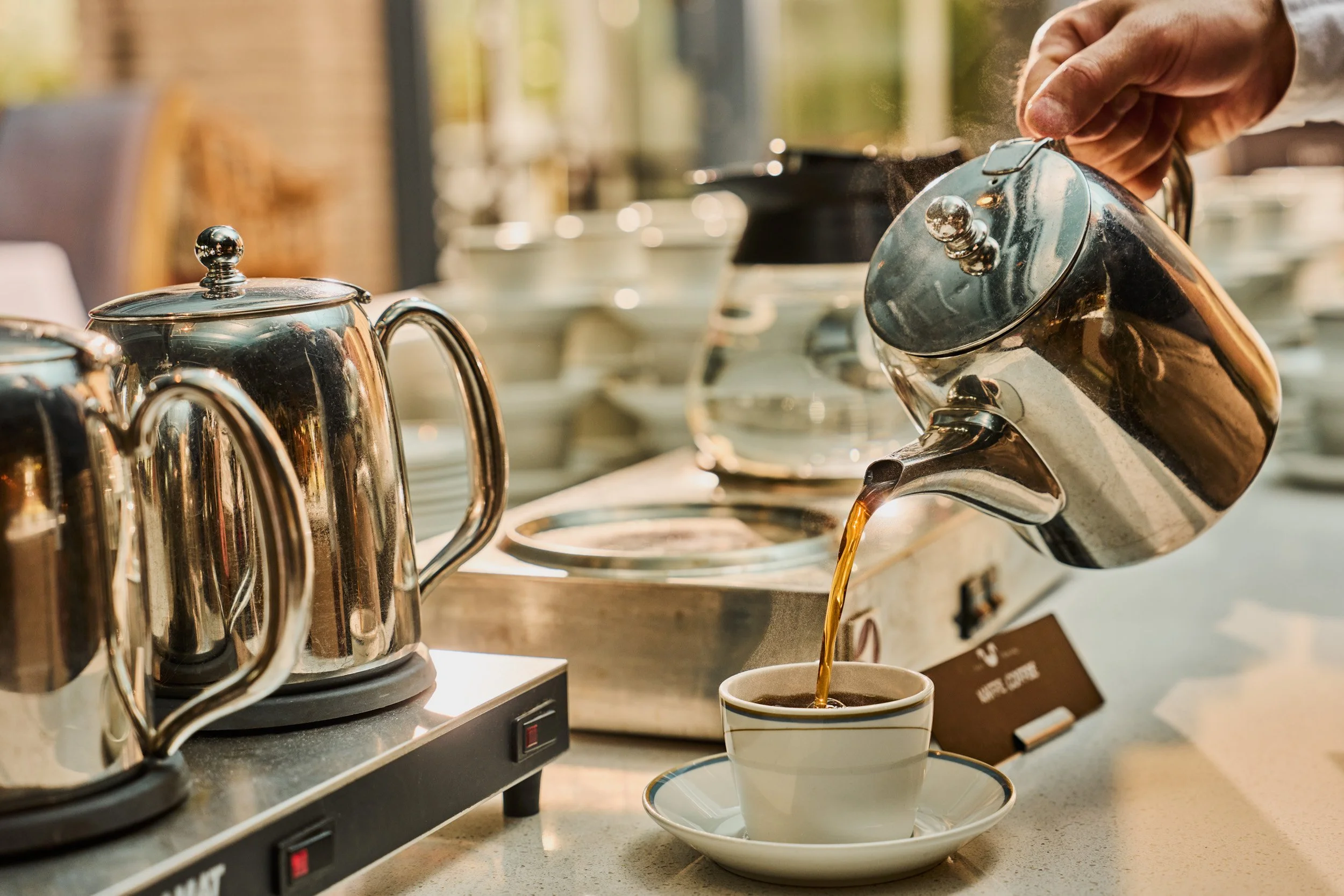 A waiter at Hotel Ullensvang pouring hot coffee from a stainless steel teapot into a cup on a saucer, with other teapots and dishes on a counter.
