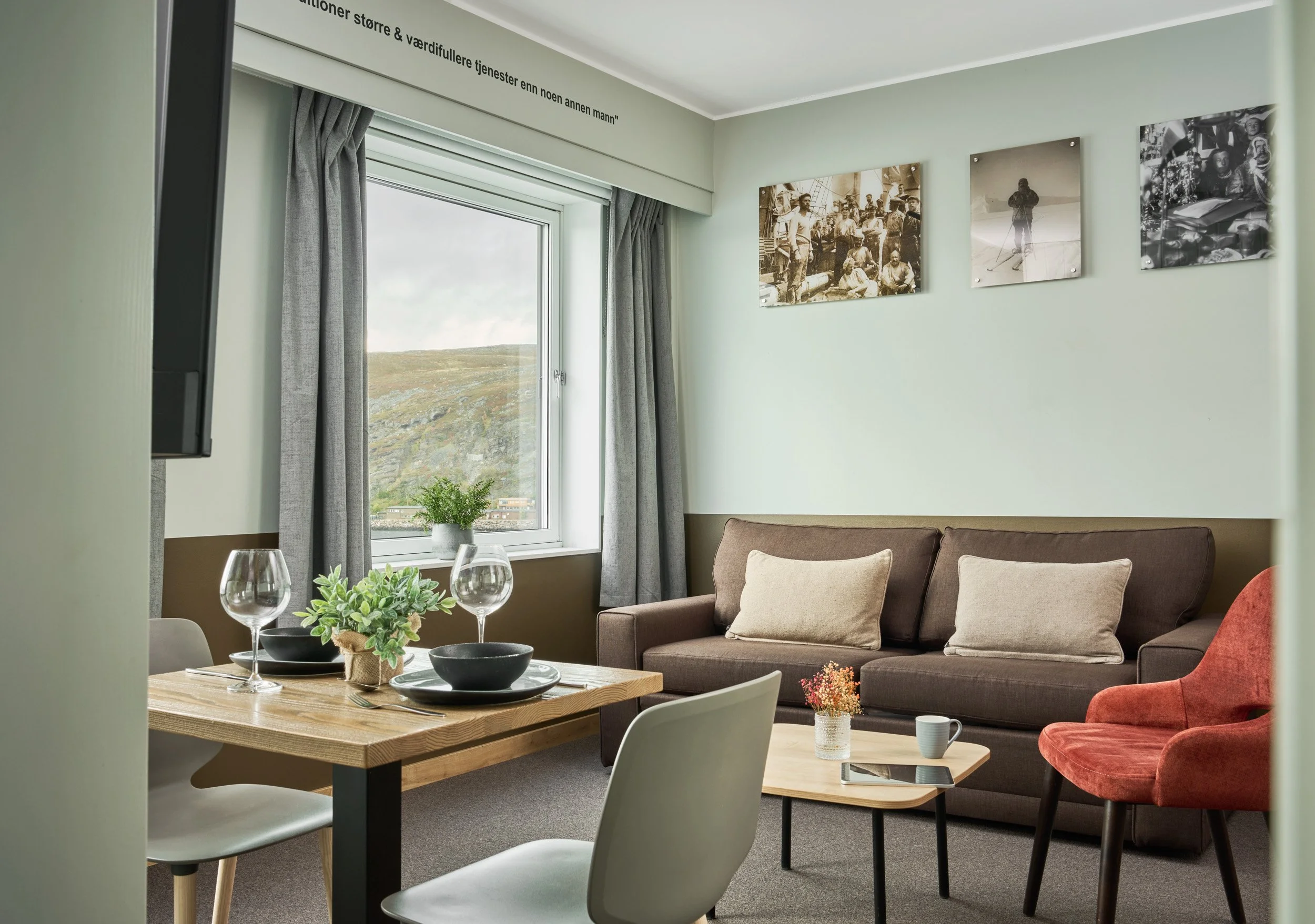 Cozy living room with a brown sofa, beige pillows, red armchair, small coffee table with flowers, dining table set with wine glasses and black bowls, window with view of a hillside, wall art, and a potted plant.