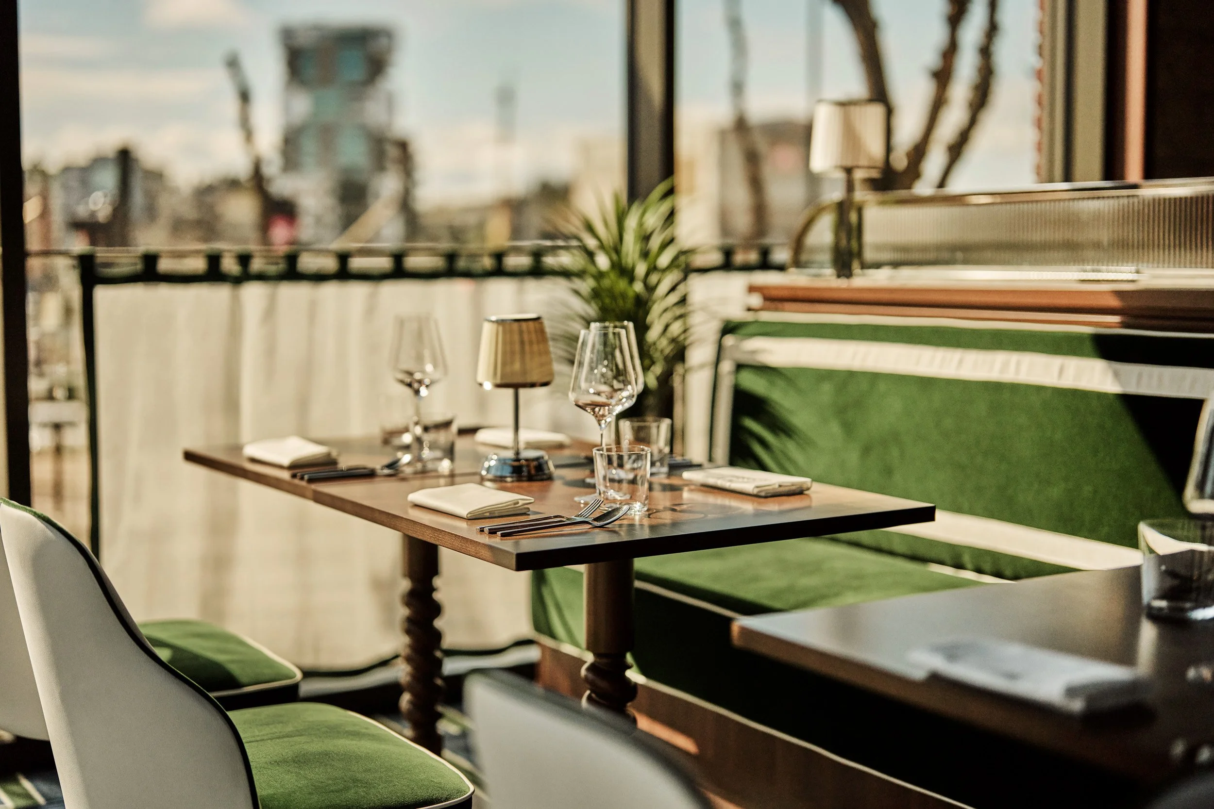 Restaurant table set with wine glasses, napkins, and cutlery, next to a window with city view and sunlight streaming in.