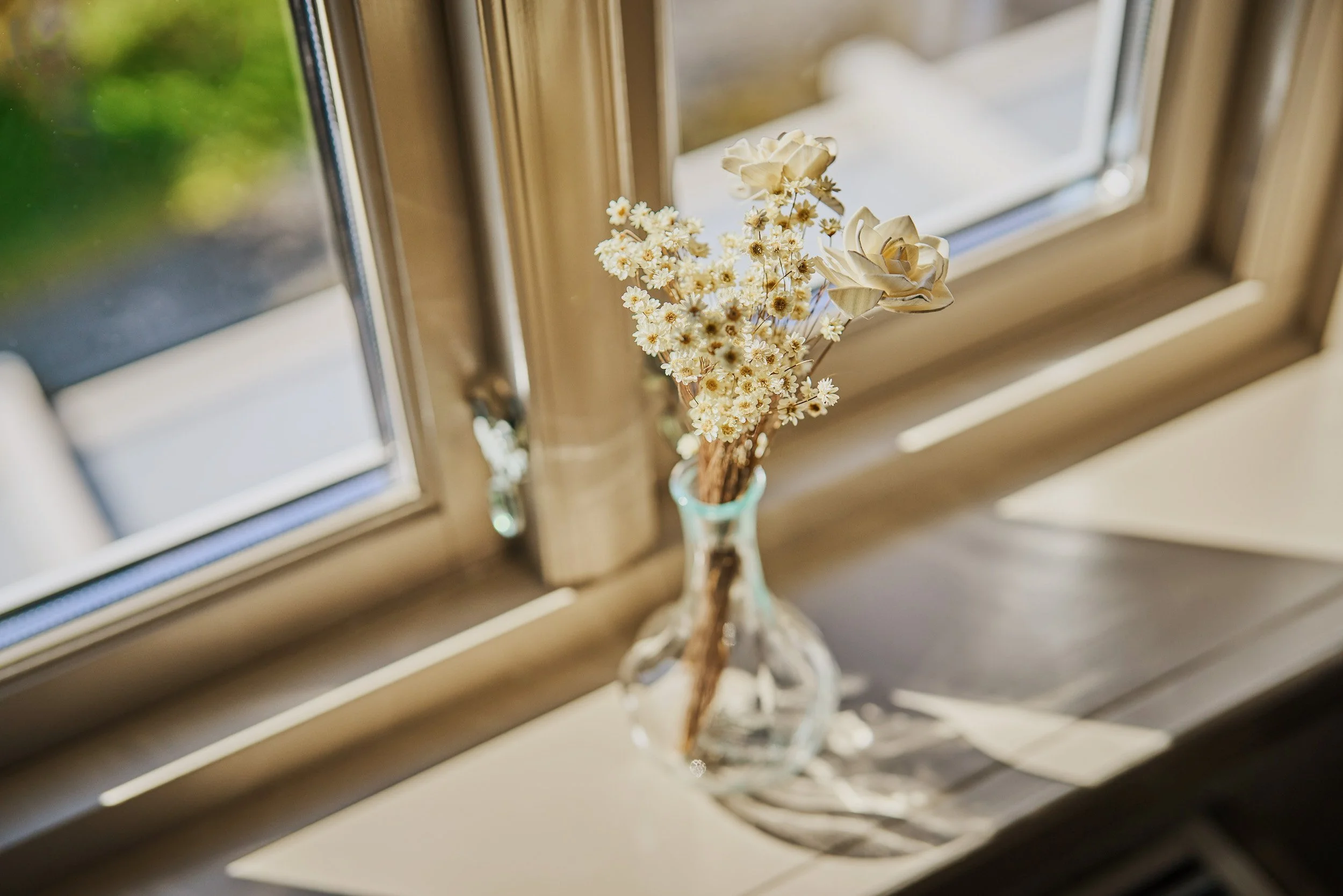 A small bouquet of white flowers in a clear glass vase on a windowsill with sunlight streaming through the window.