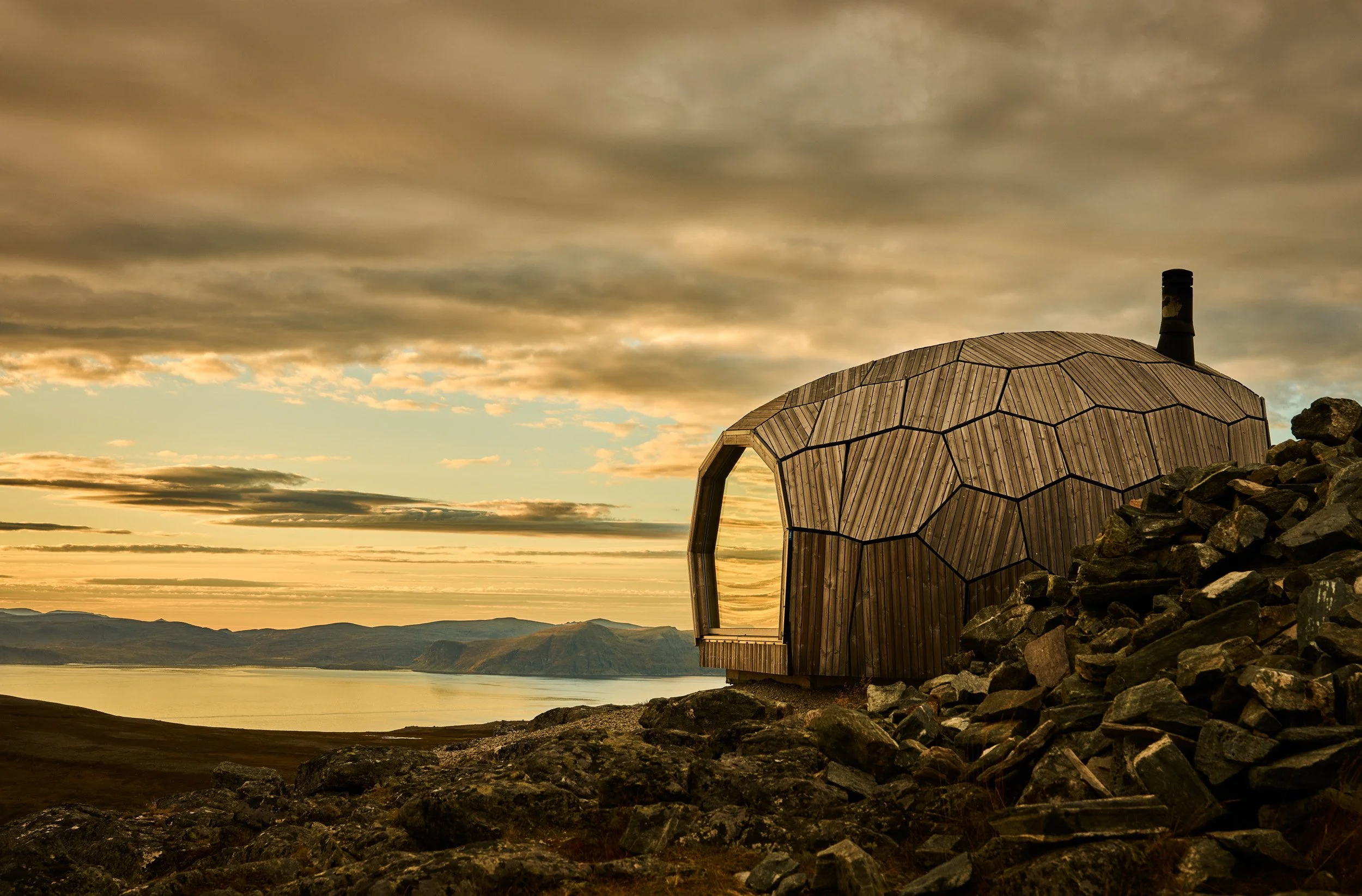 A unique wooden building with a dome-shaped structure and honeycomb pattern, situated on rocky terrain overlooking a body of water at sunset.