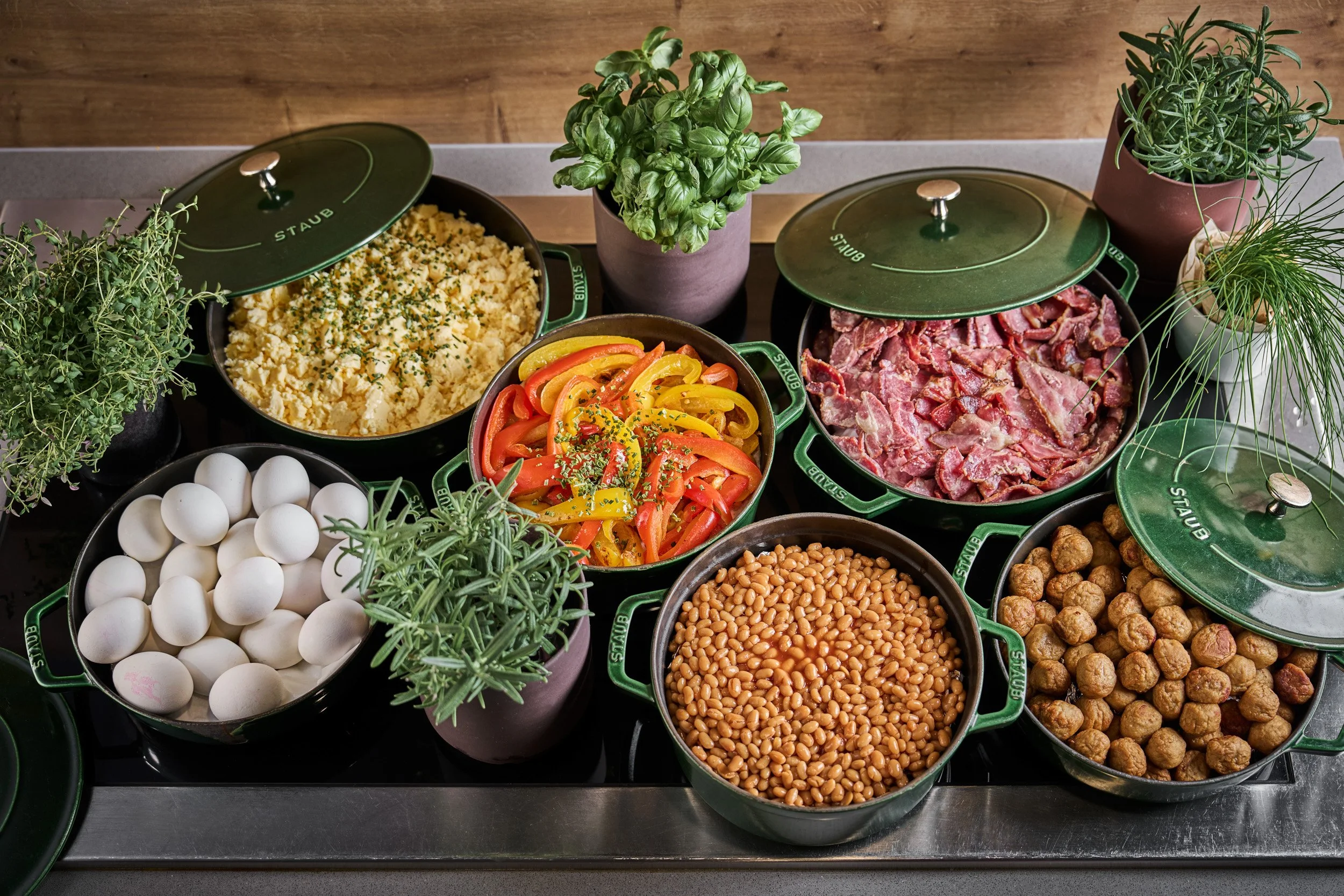 Various ingredients in green Staub pots and bowls, including eggs, chopped bacon, roasted chickpeas, colorful sliced bell peppers, herbs, and cauliflower mash, arranged on a kitchen countertop.