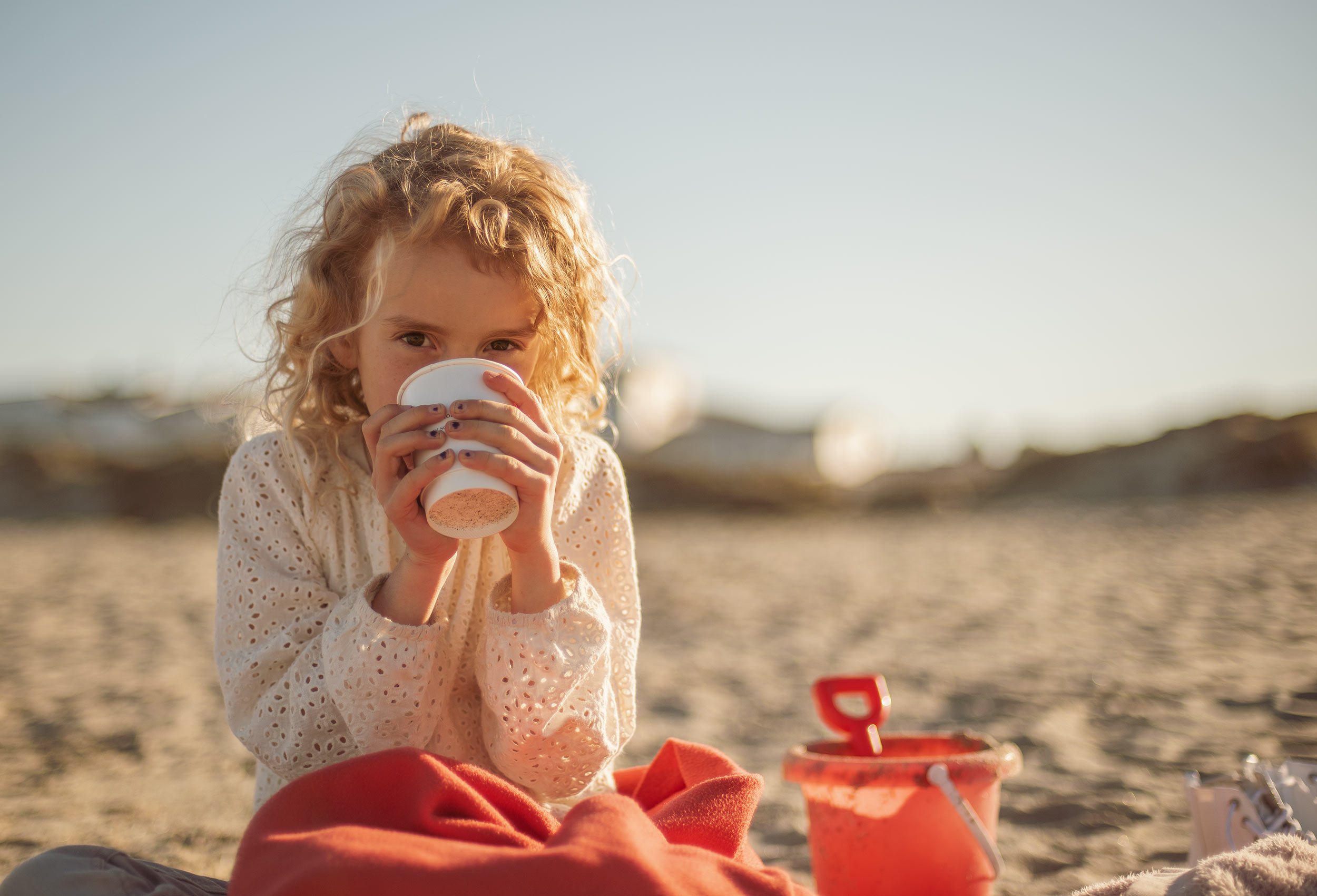 girl playing with a red pail and spade on the Sola beach and drinking cocoa in the sunset