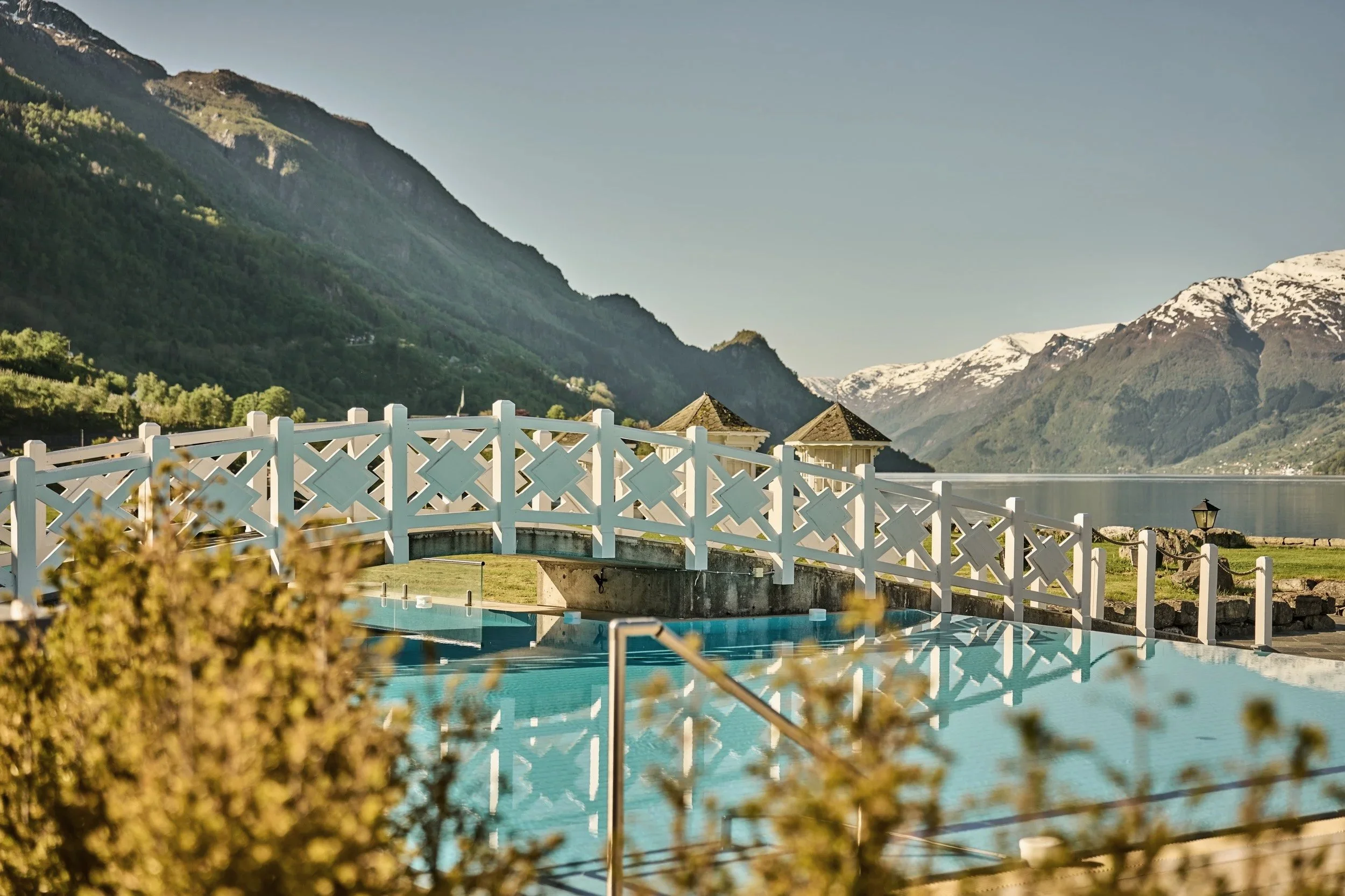 A scenic view of a swimming pool with a white fence, overlooking a lake surrounded by mountains with snow-capped peaks in the background.
