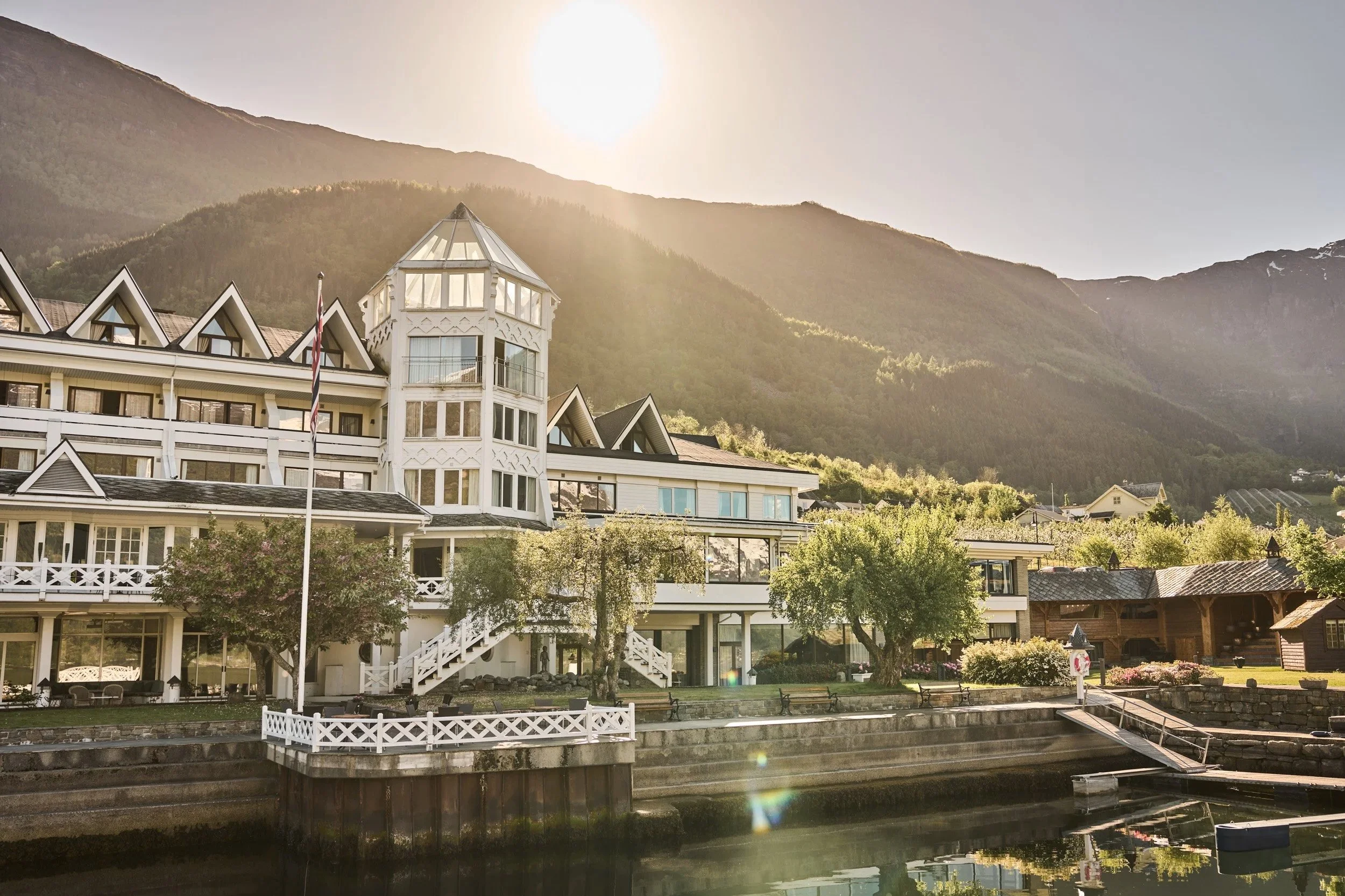A large, white hotel with a tower, situated by a body of water with steps leading down to the water, surrounded by trees and mountains in the background, with the sun shining.