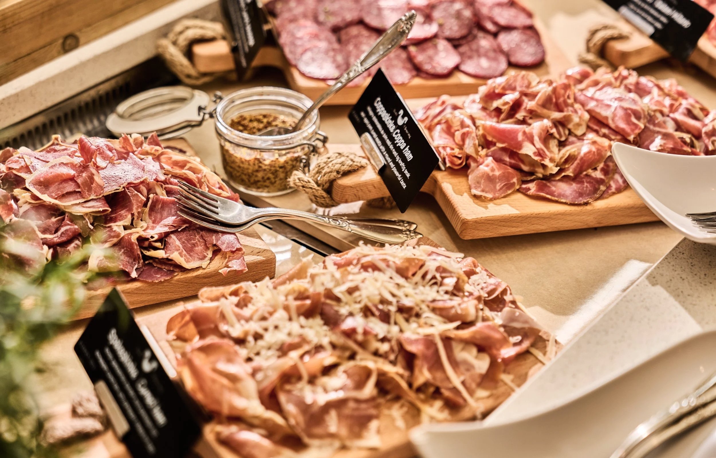 An assortment of cured meats and charcuterie on wooden boards, including sliced ham, salami, and prosciutto, with small bowls of mustard and condiments.