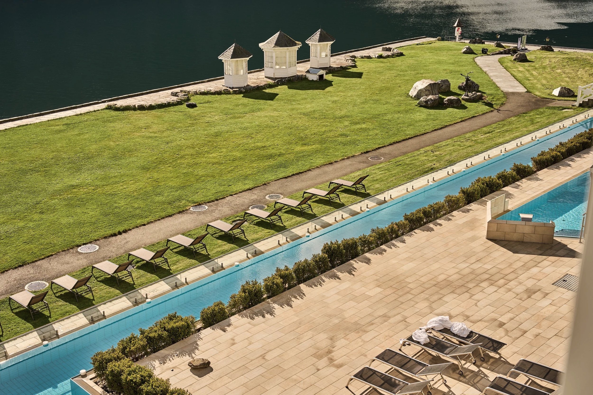 Poolside area at Hotel Ullensvang with lounge chairs, towels, and cushions, adjacent to a garden with a grassy lawn and a pathway, next to a body of water with small gazebos and rocks.