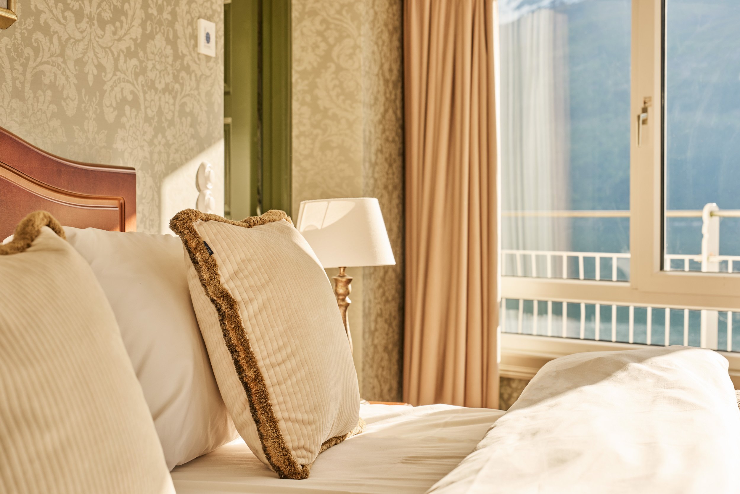 Cozy bed with cream-colored pillows and a bedside lamp, sunlight streaming through a window with open curtains in a warmly lit bedroom.