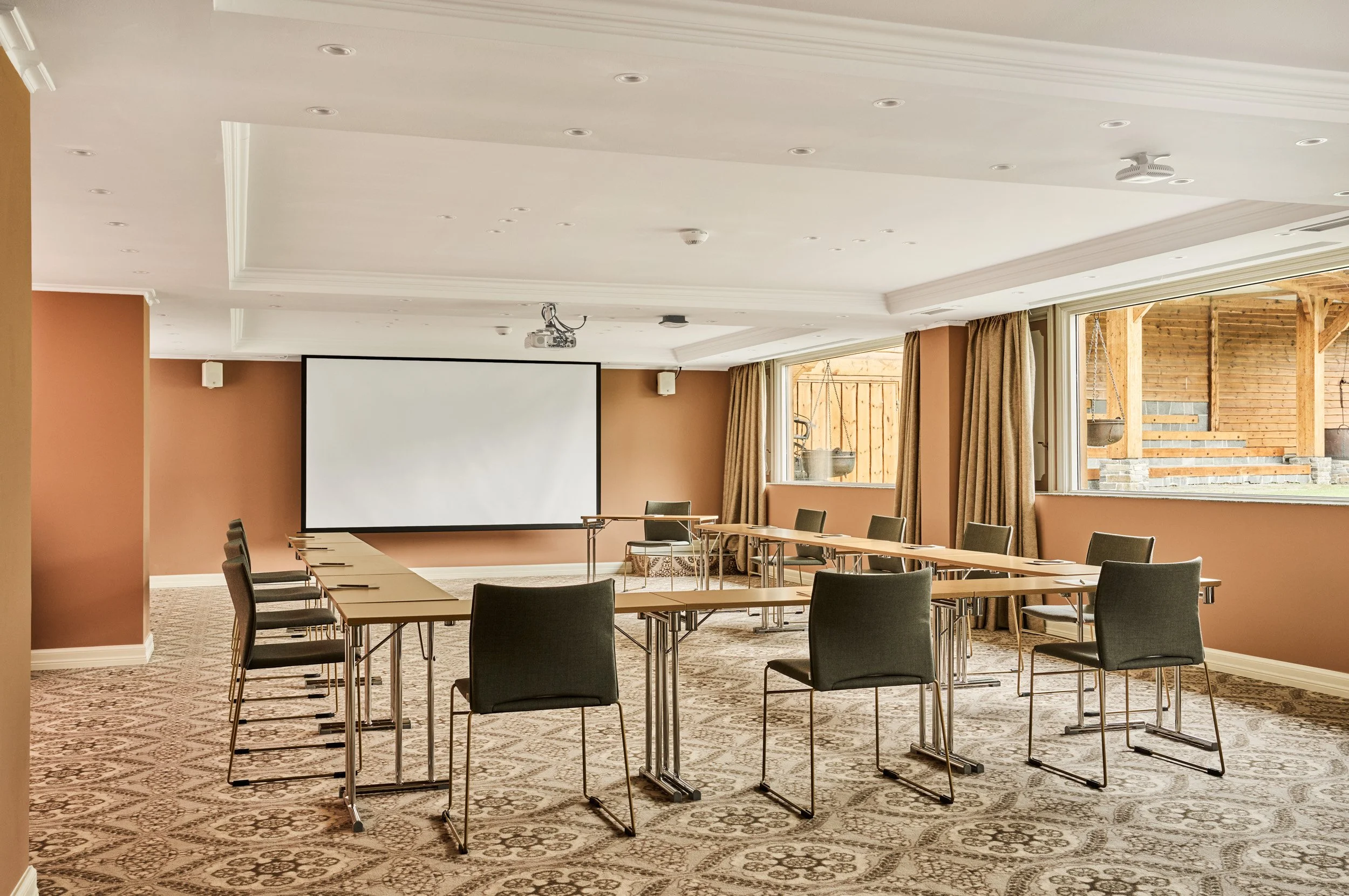 Empty conference room with U-shaped arrangement of tables and chairs, a large screen on the wall, large windows with curtains, and a patterned carpet.