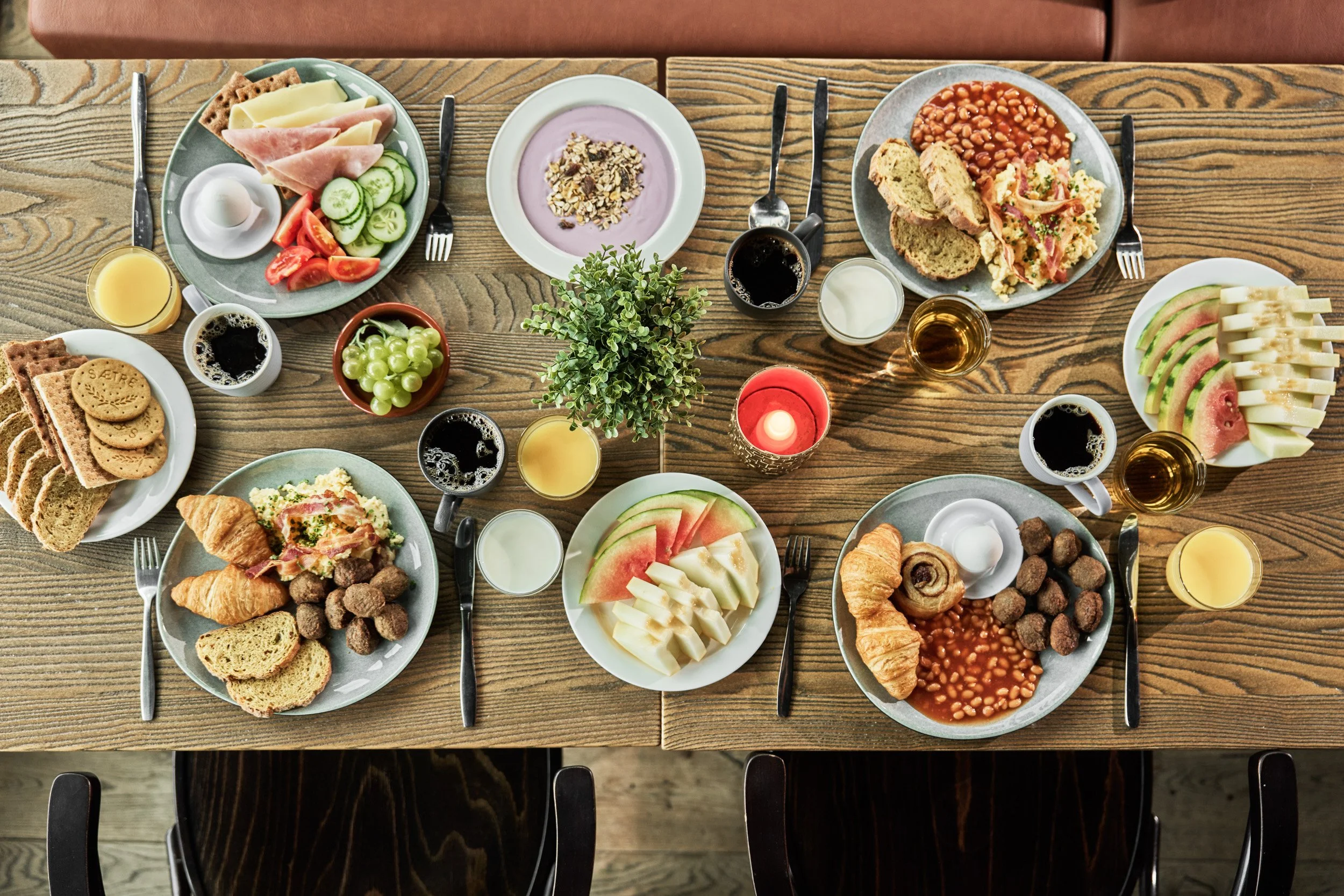 A large wooden table set for a breakfast, with plates of mixed fruits, pastries, bread, cheese, meats, yogurt, and various beverages including coffee, juice, and water, decorated with small candles and a plant in the center.