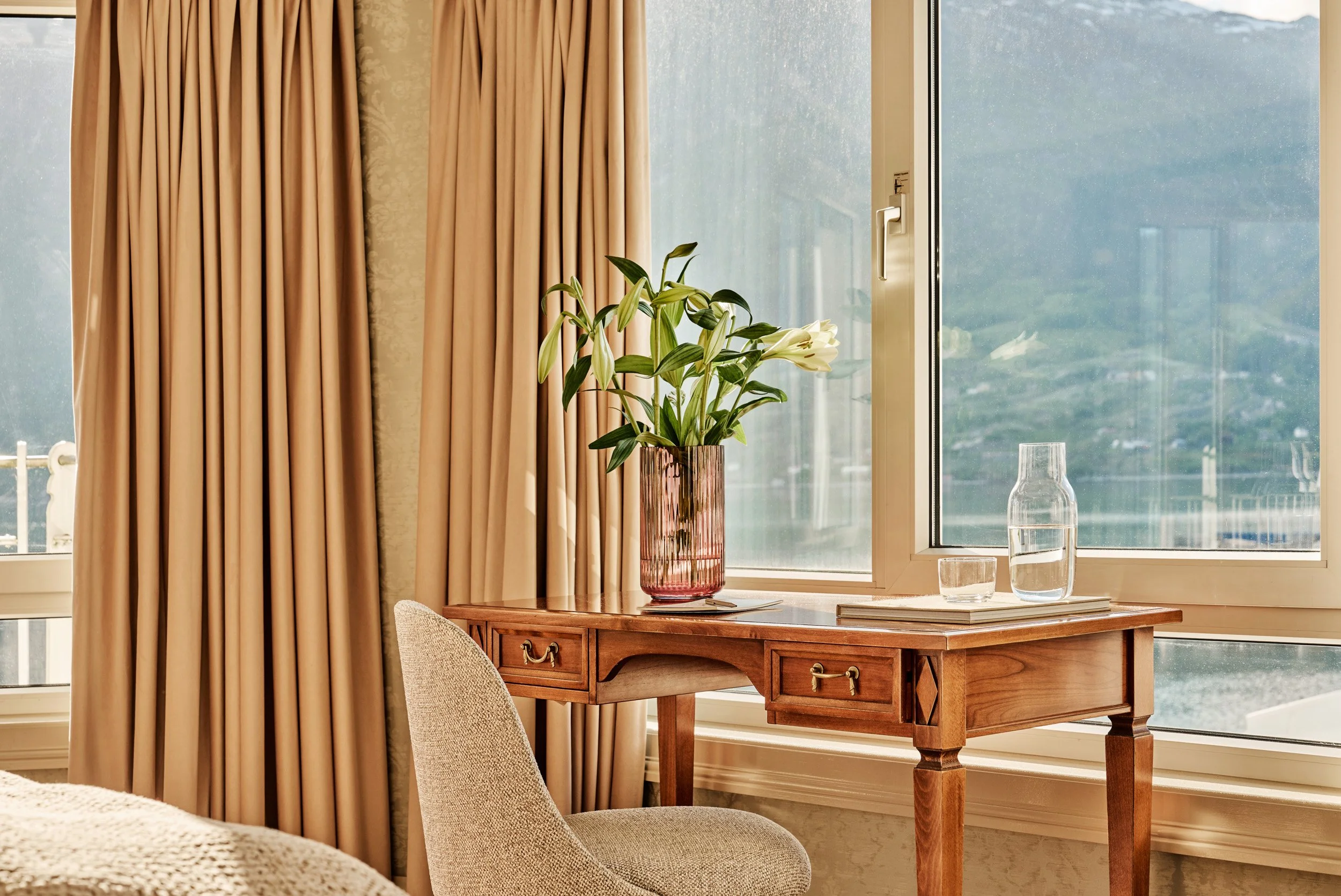 A wooden desk with a pink glass vase holding white lilies, a glass water pitcher, and a glass of water by a window with beige curtains.