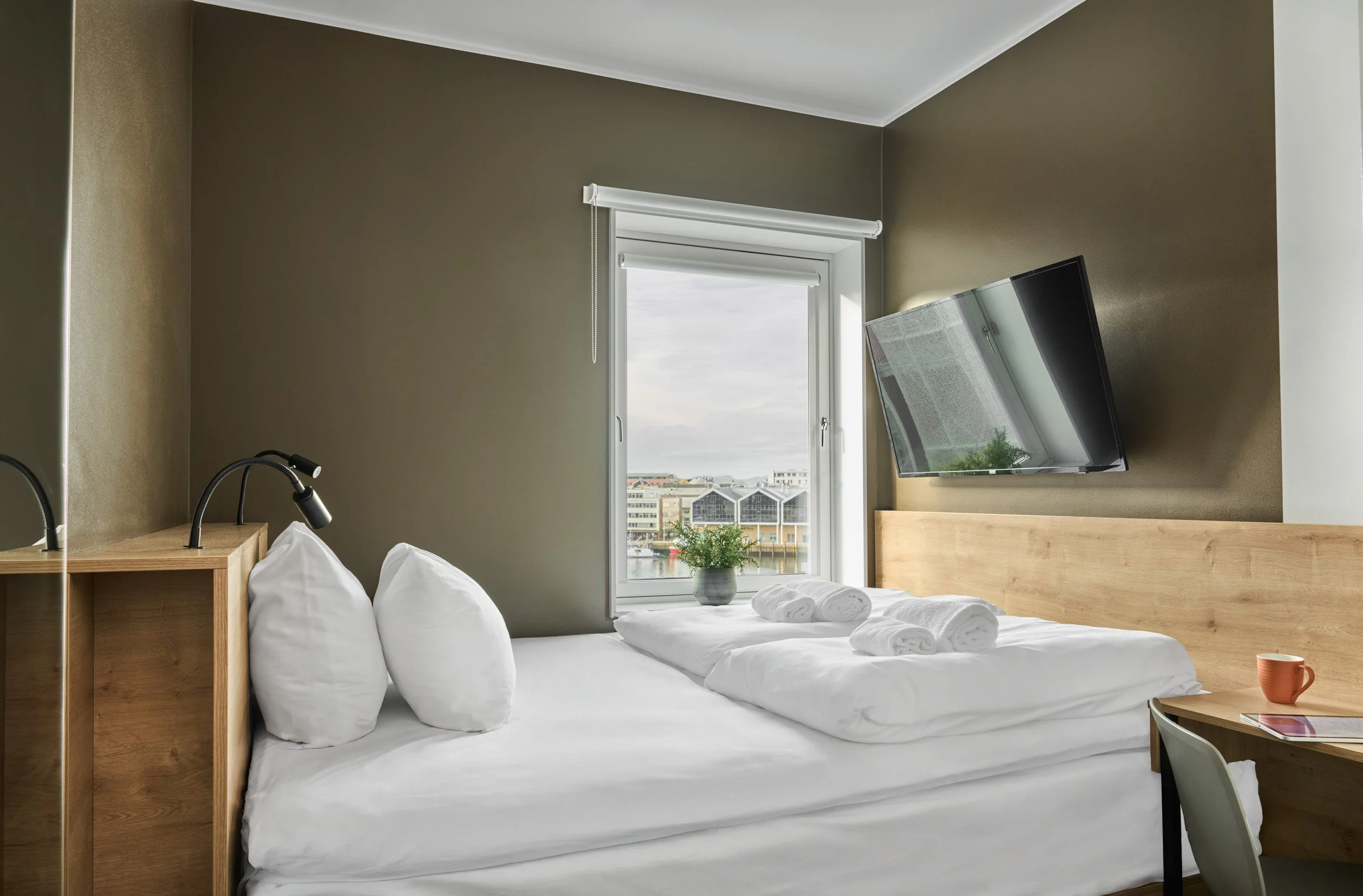 A modern hotel room with a double bed, white pillows, and rolled towels, a window with a view of city buildings, a wooden headboard, a wall-mounted flat-screen TV, and a small wooden desk with a coffee mug and magazine.
