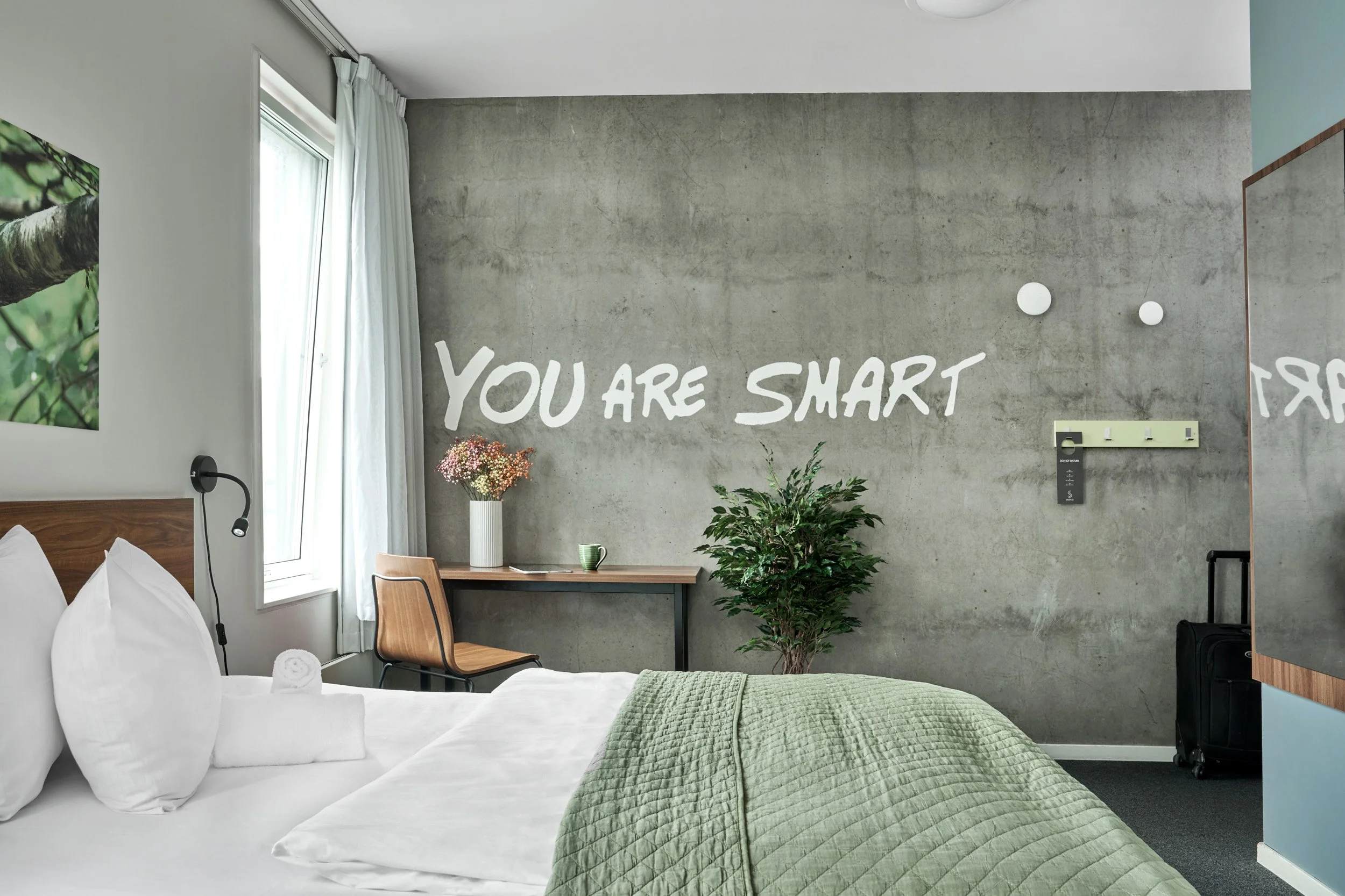 A hotel room with a bed, a window with white curtains, a desk with a vase of flowers and a green cup, a chair, a plant, and a wall with the message "You are smart" painted on it.