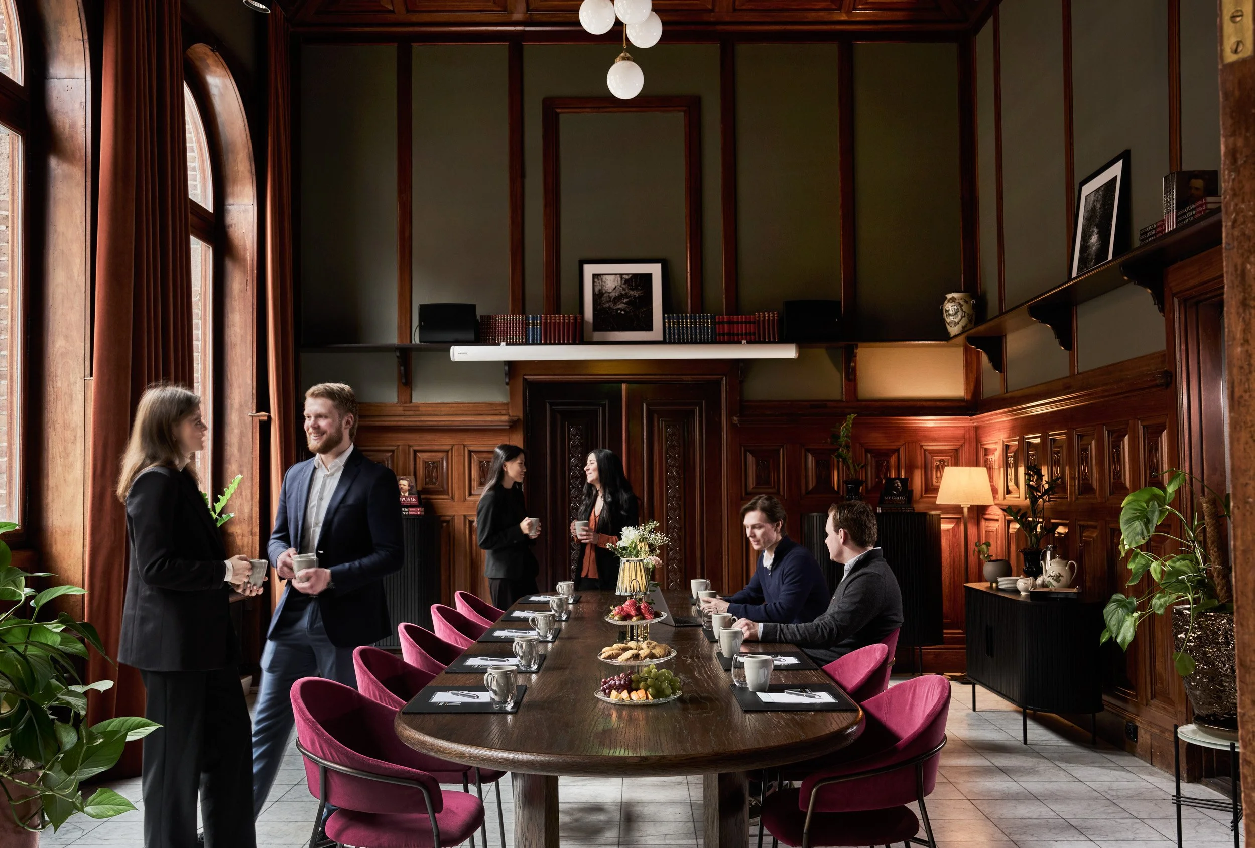 Business meeting in a formal conference room with six people, including three men and three women, engaging in conversations and enjoying refreshments around a long wooden table with pink chairs. The room features wood-paneled walls, large windows wi