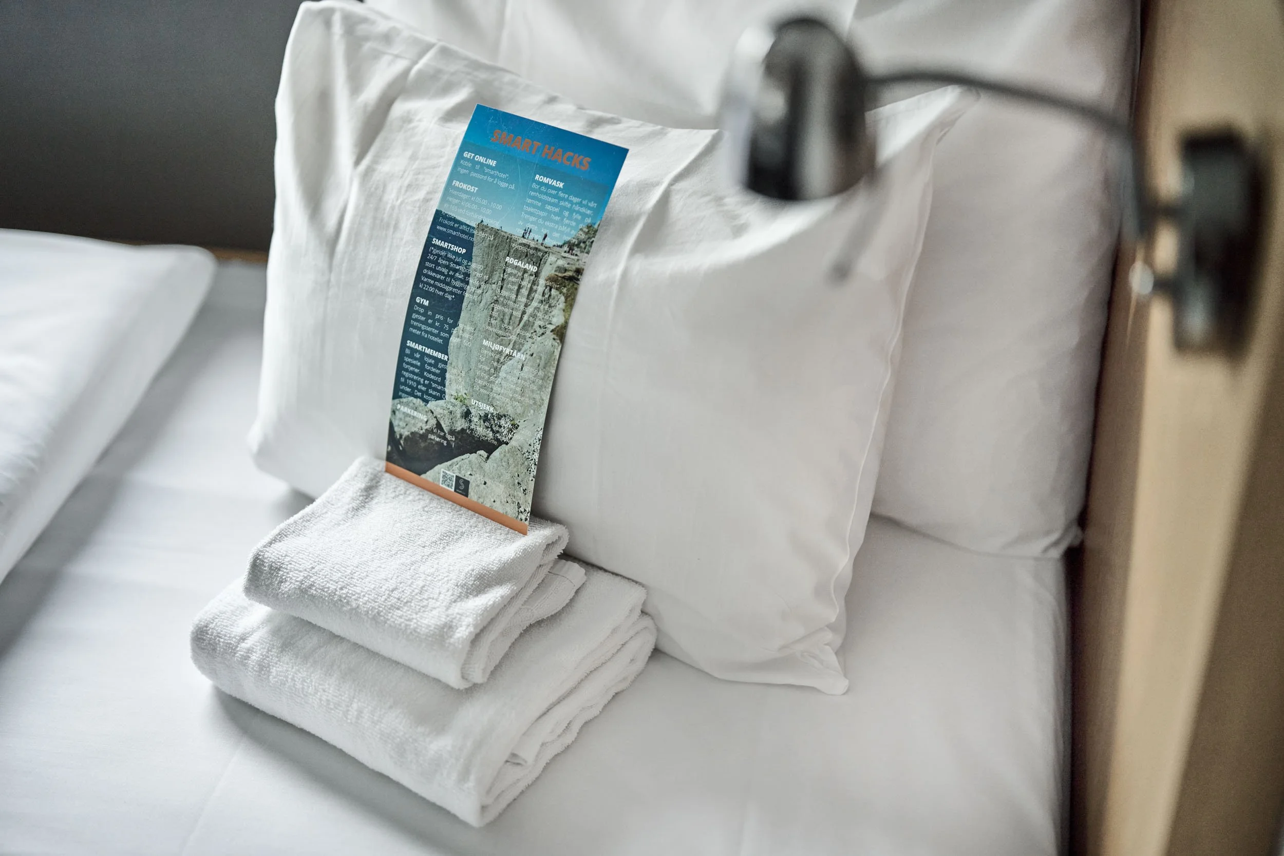 A neatly made bed with white pillows, a folded white towel, and a colorful informational brochure standing against the pillows.