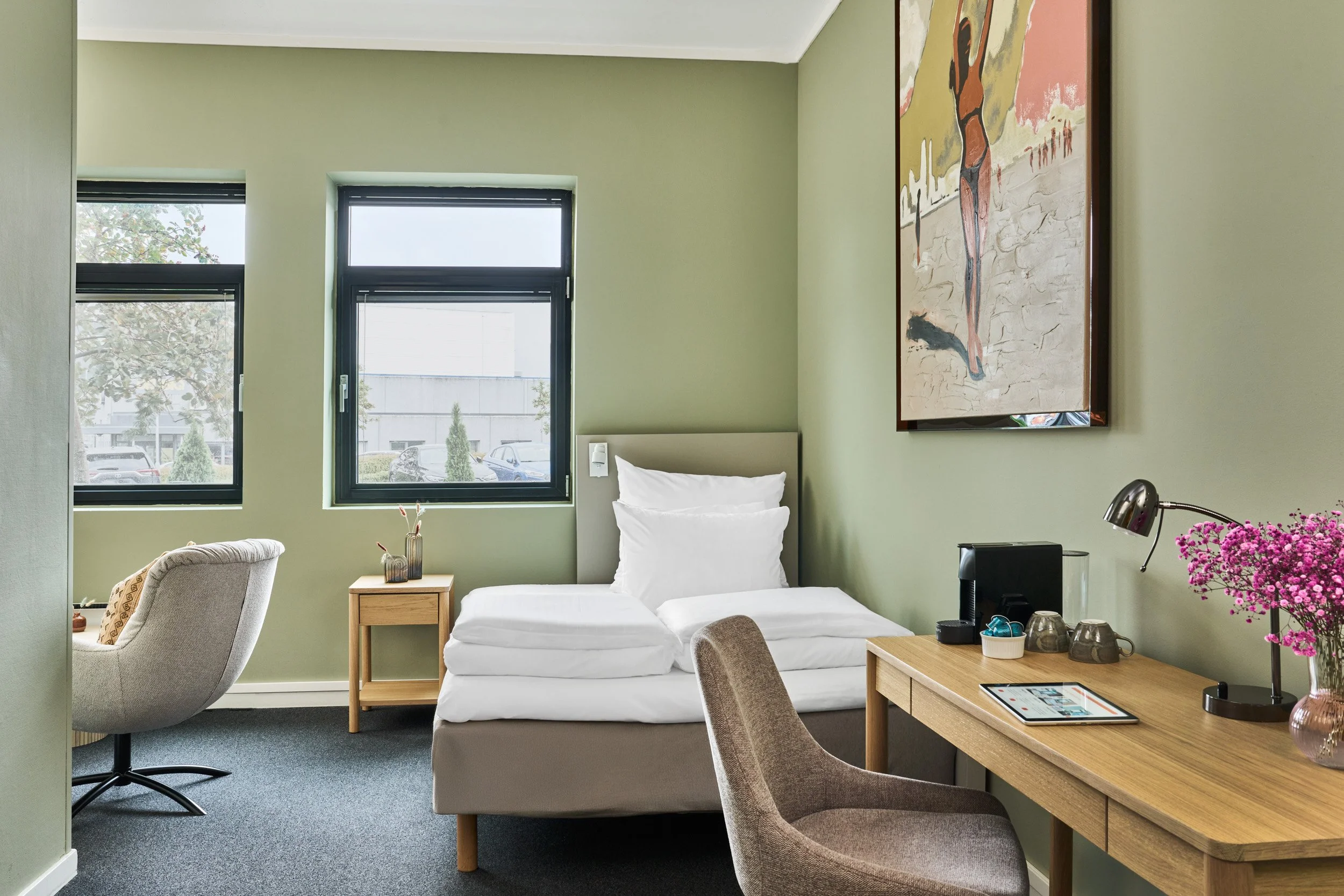 A hotel room with green walls, two windows, an unmade bed with white bedding, a wooden desk with a lamp, flowers, and a tablet, a gray chair, and artwork on the wall.