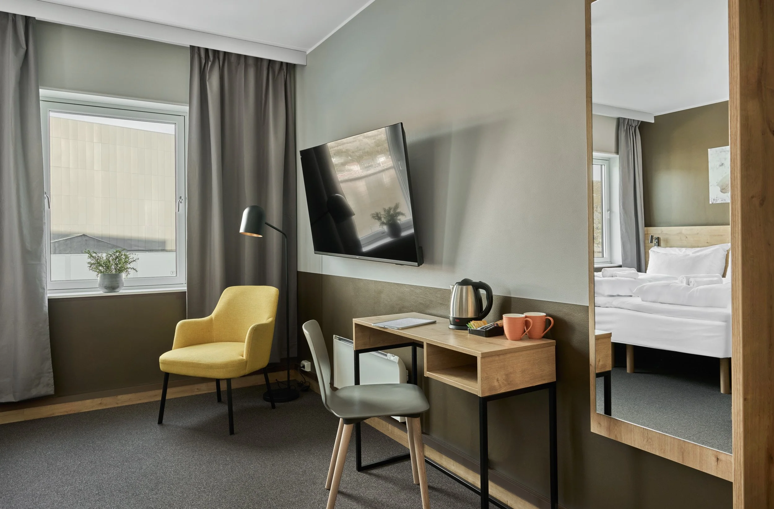 Hotel room with a window, yellow armchair, flat-screen TV, small desk with kettle and mugs, mirror, and a partial view of a bedroom with bed and nightstand.