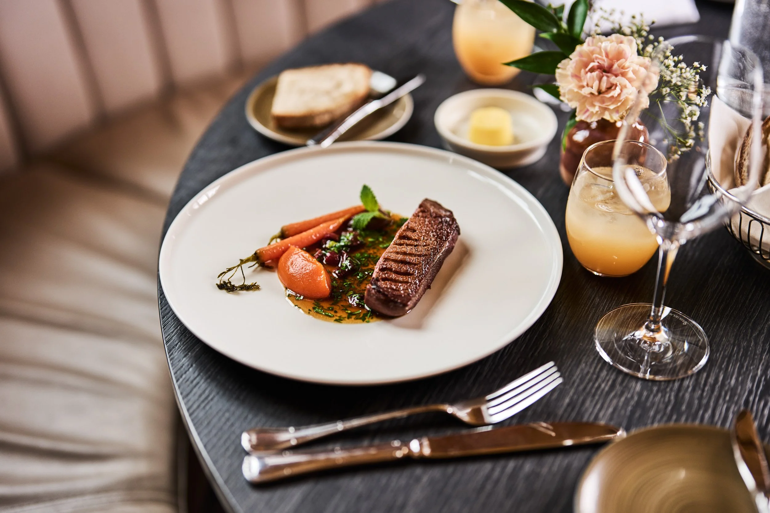 A plate with a cooked piece of beef, carrots, and sauce on a dining table, surrounded by wine glasses, a fork and knife, a small dish with butter, a slice of bread, and flowers.