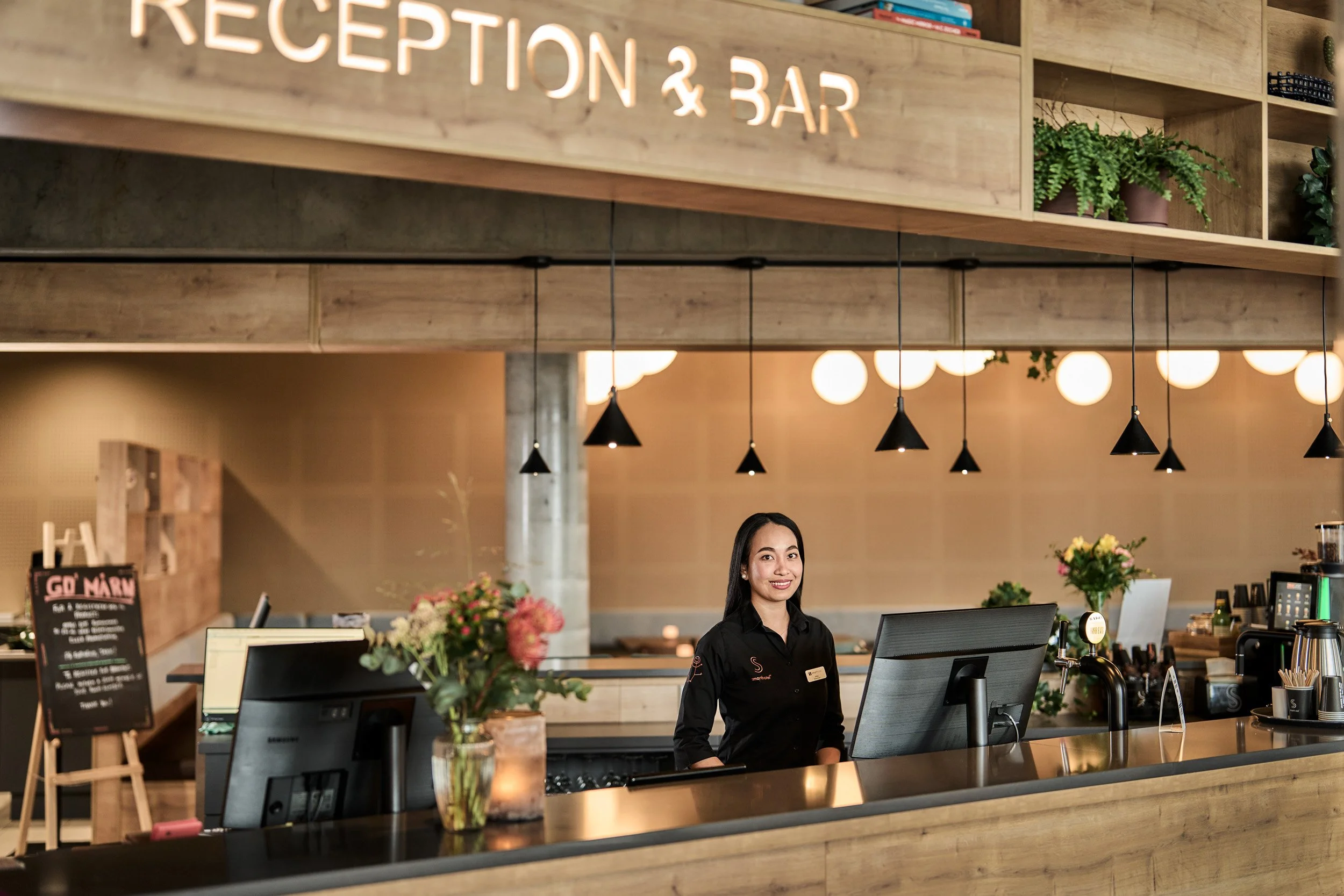 Reception area at a hotel with a smiling female staff member standing behind the counter, decorated with flowers and digital screens, illuminated by black pendant lights.