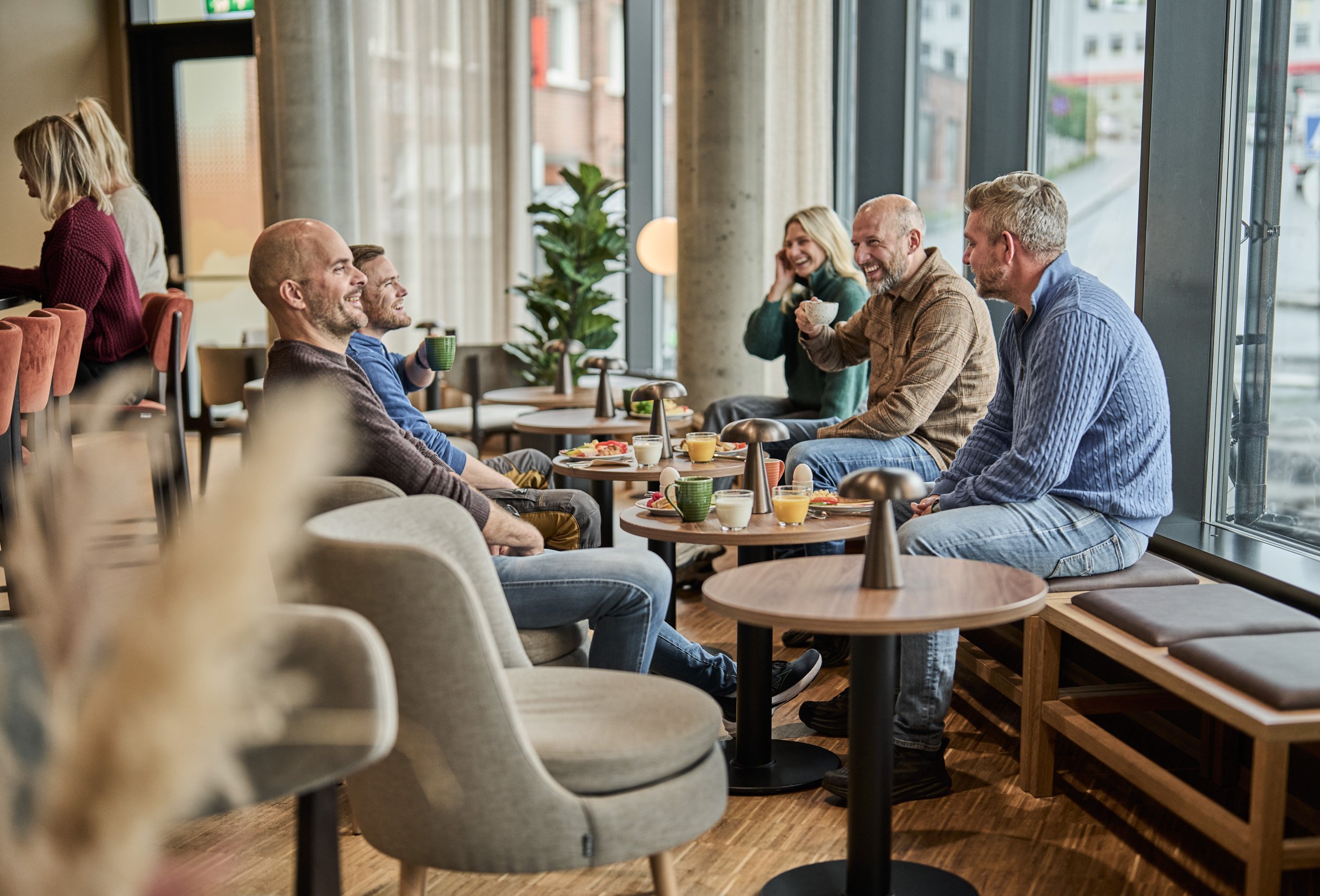 Group of six people sitting around small tables in a cafe, enjoying coffee and snacks, smiling and engaging in conversation, with large windows letting in natural light.