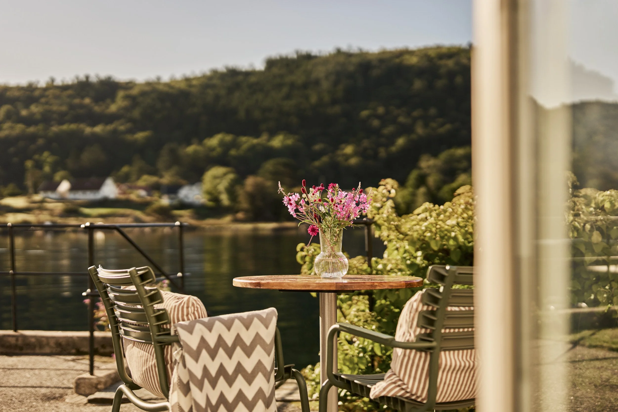 Outdoor patio table with pink flowers in a glass vase, surrounded by metal chairs with striped cushions, overlooking a river with a wooded hillside in the background.