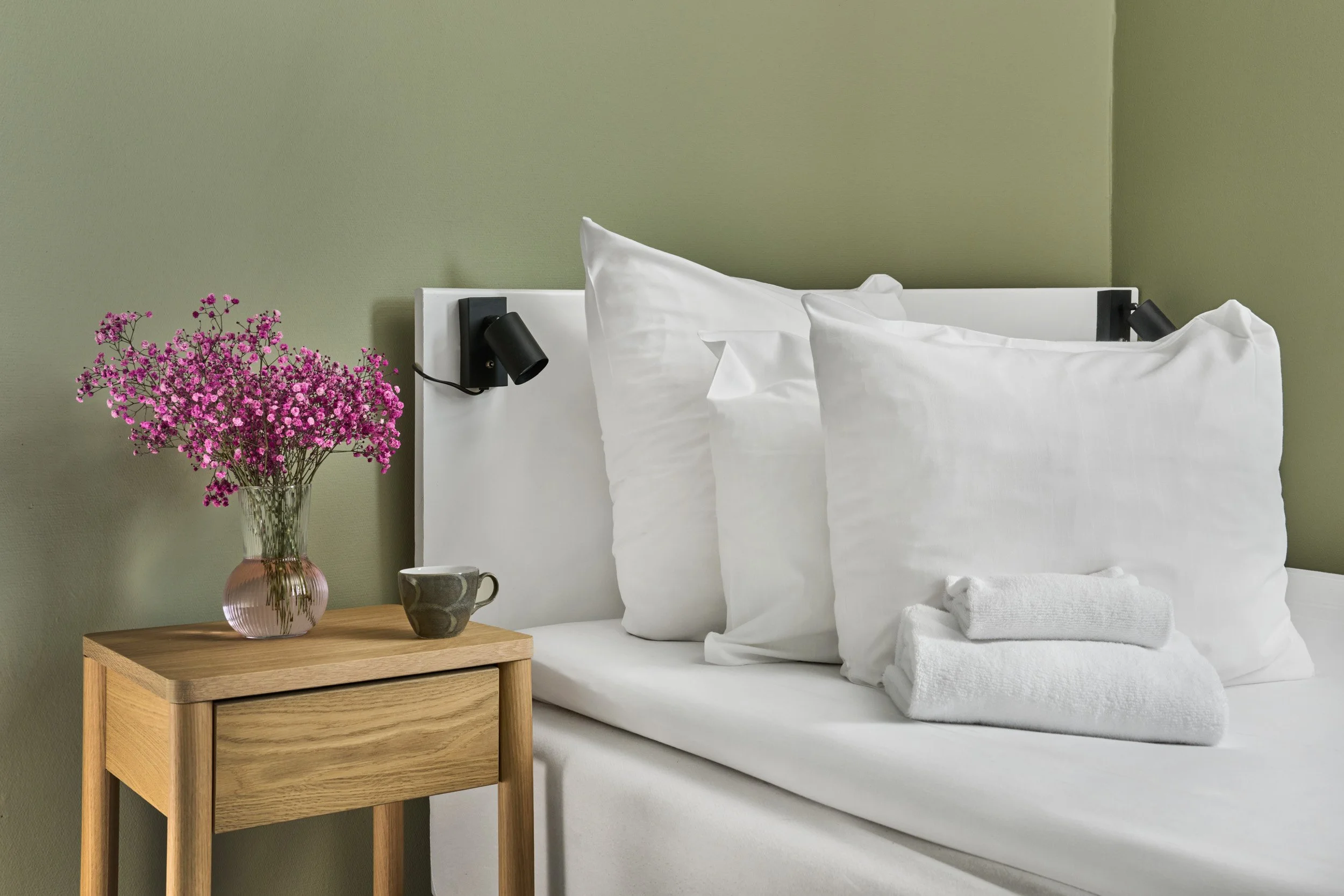 A bedroom with a bed, white linens, oversized white pillows, a bedside table with a vase of pink flowers, a coffee mug, and folded white towels, against a light green wall with black reading lights.