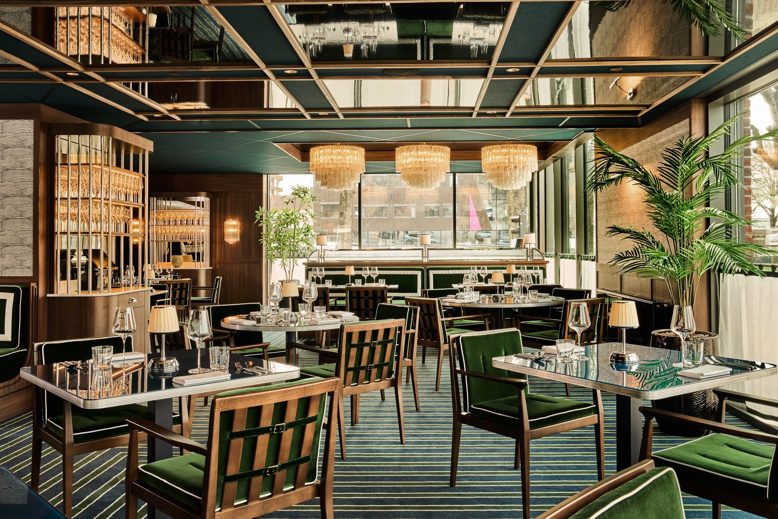Elegant restaurant interior with dark wood furniture, green upholstered chairs, chandeliers, large windows, potted plants, and a striped carpet.