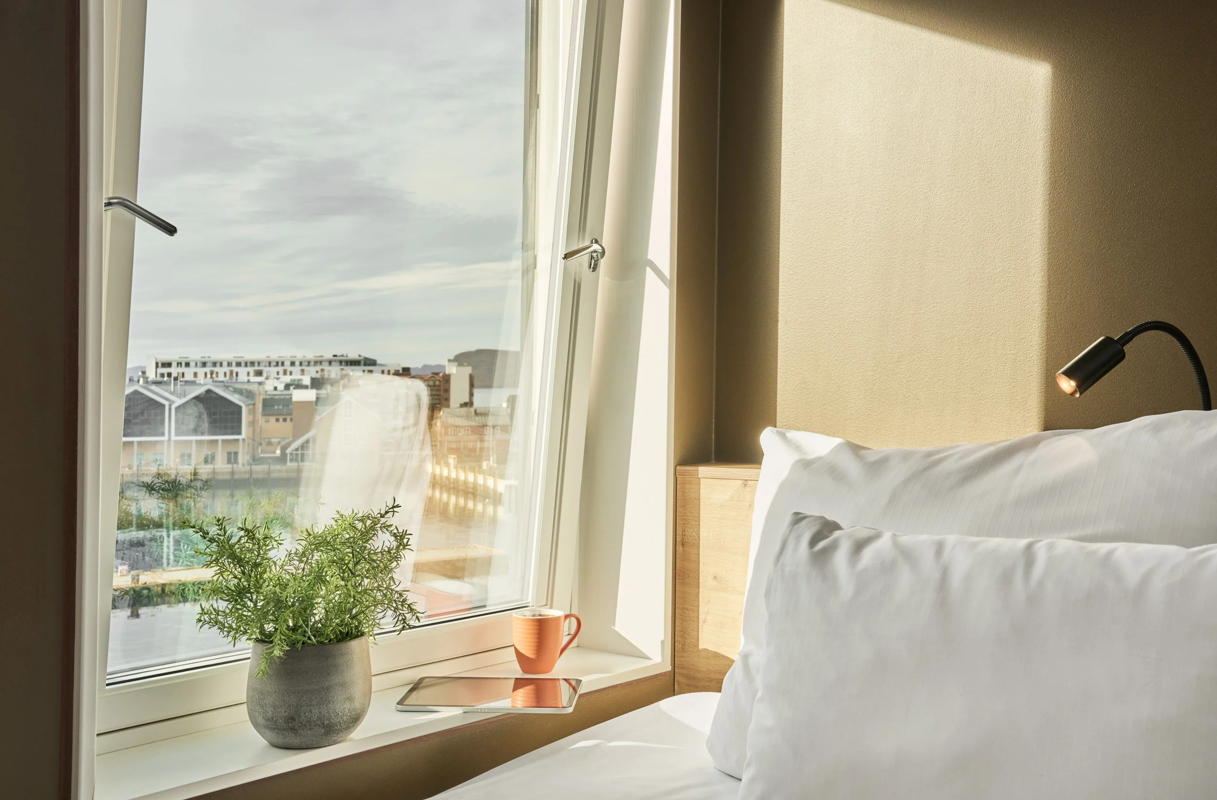 Sunlit hotel room with a bed, white pillows, a black adjustable reading lamp, a potted plant, a tablet, an orange mug, and an open window showing a cityscape and cloudy sky.