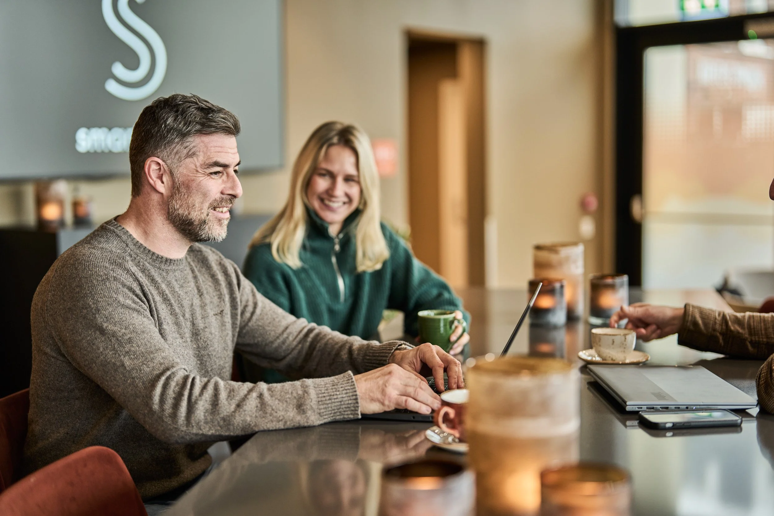 A group of people sitting at a coffee shop table, engaging in conversation and smiling, with laptops, cups, and candles on the table.