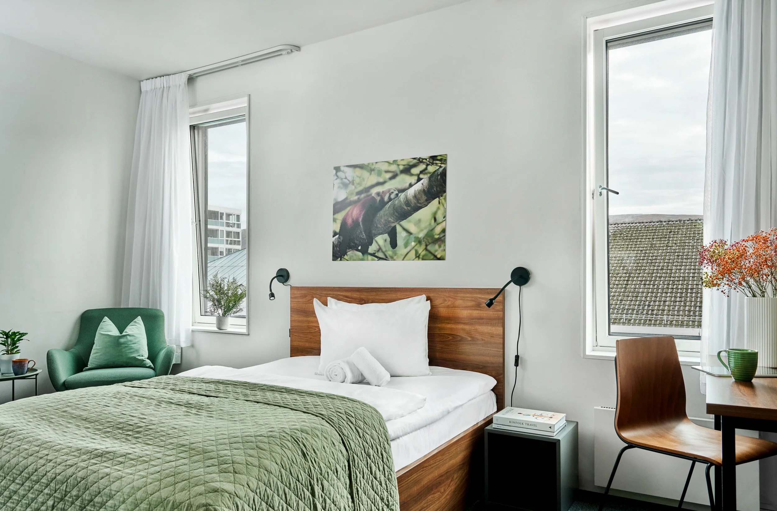 Bedroom with white walls, wooden bed with white bedding, green quilt, and pillows. Art featuring a bird on a branch hangs above the bed. A green armchair with a pillow is in the corner, next to a small table with plants. A desk with a chair and flowe