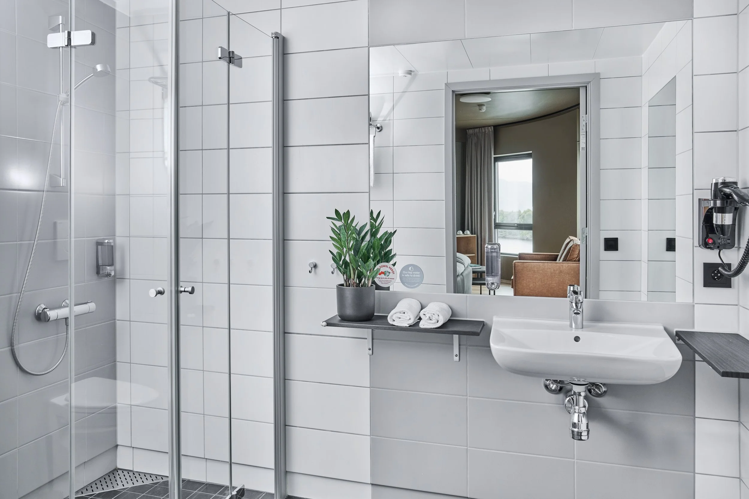 Modern bathroom with white tiled walls, shower area with glass door and handheld showerhead, wall-mounted sink, mirror, potted plant, and rolled towels. Through the mirror, a sitting area with a window is visible.