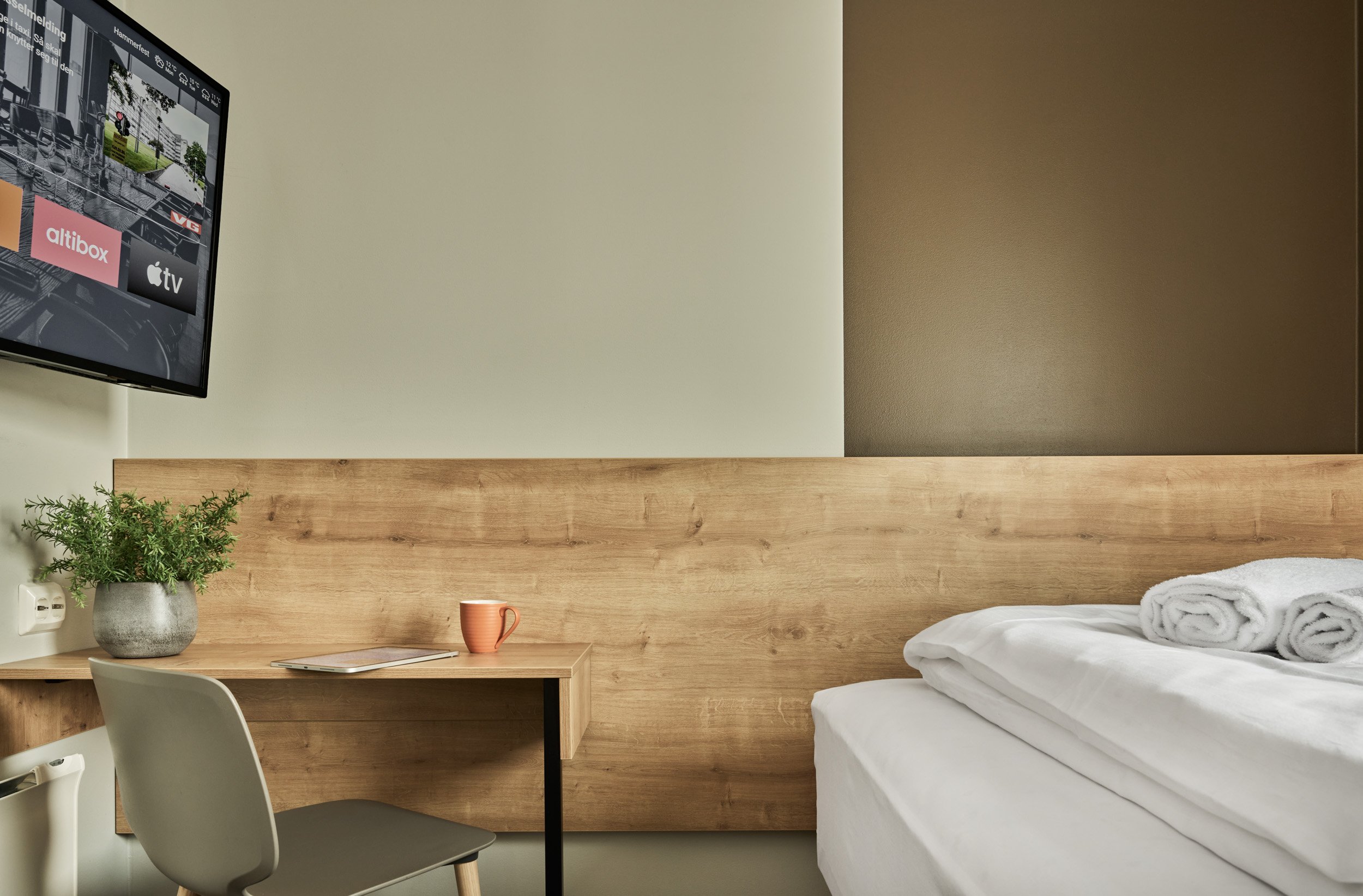 Hotel room with a bed, rolled towels, a wooden headboard, a desk with a potted plant, a beige chair, a coffee mug, and a wall-mounted TV.