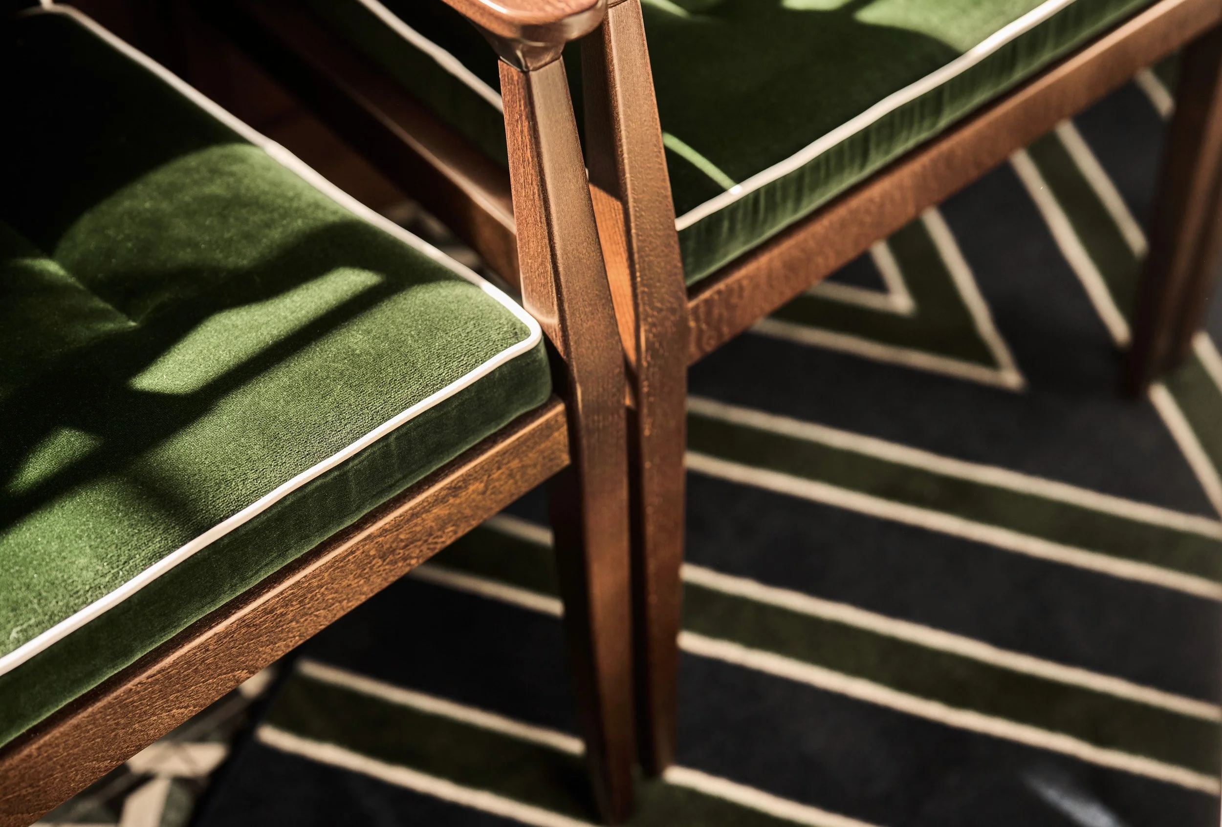 Close-up of vintage wooden chairs with green velvet cushions and white piping, showing shadows cast on the cushions and patterned carpet underneath.