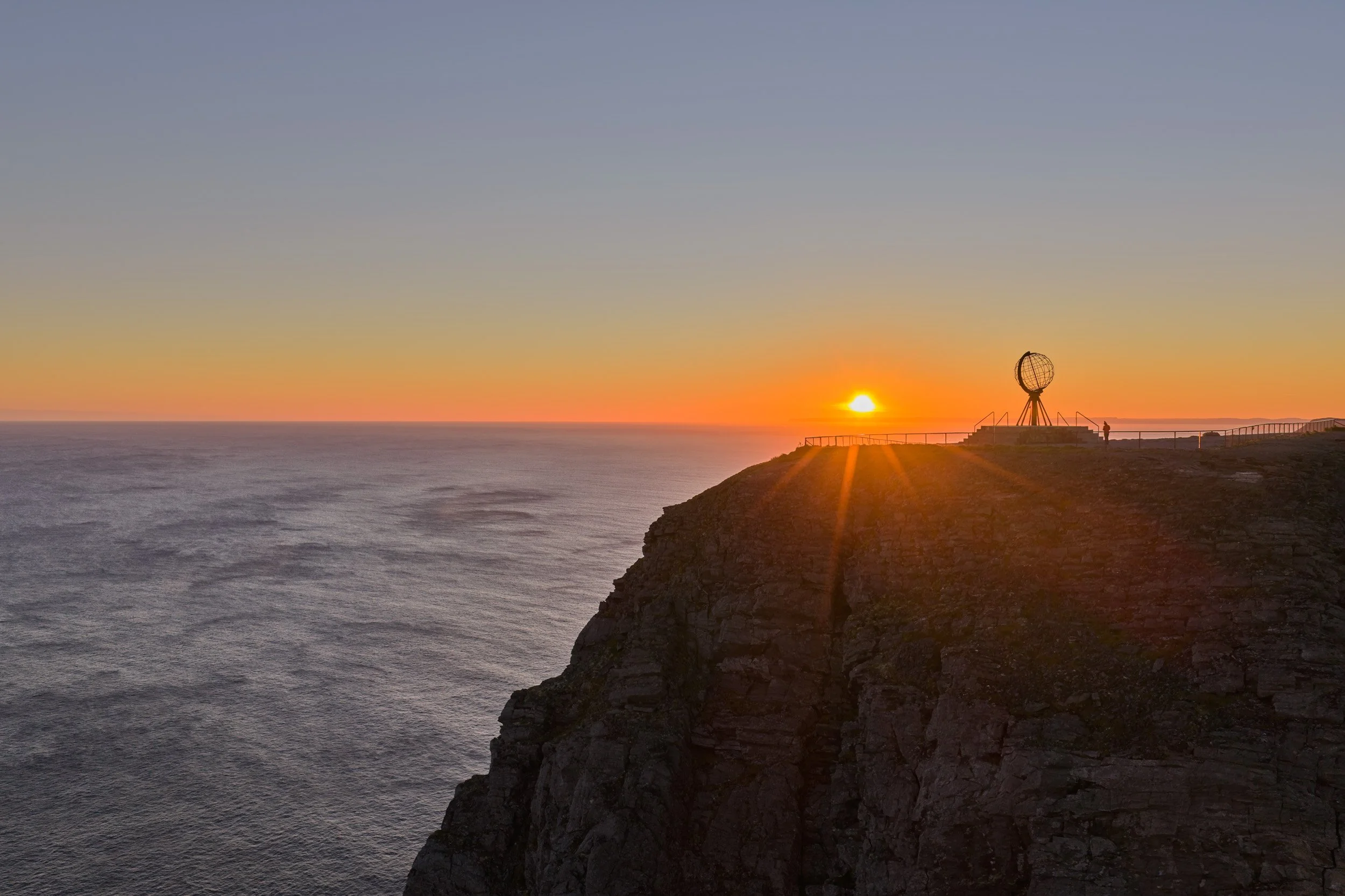 Sunset over the ocean viewed from a cliff with a spherical monument and a person standing nearby.