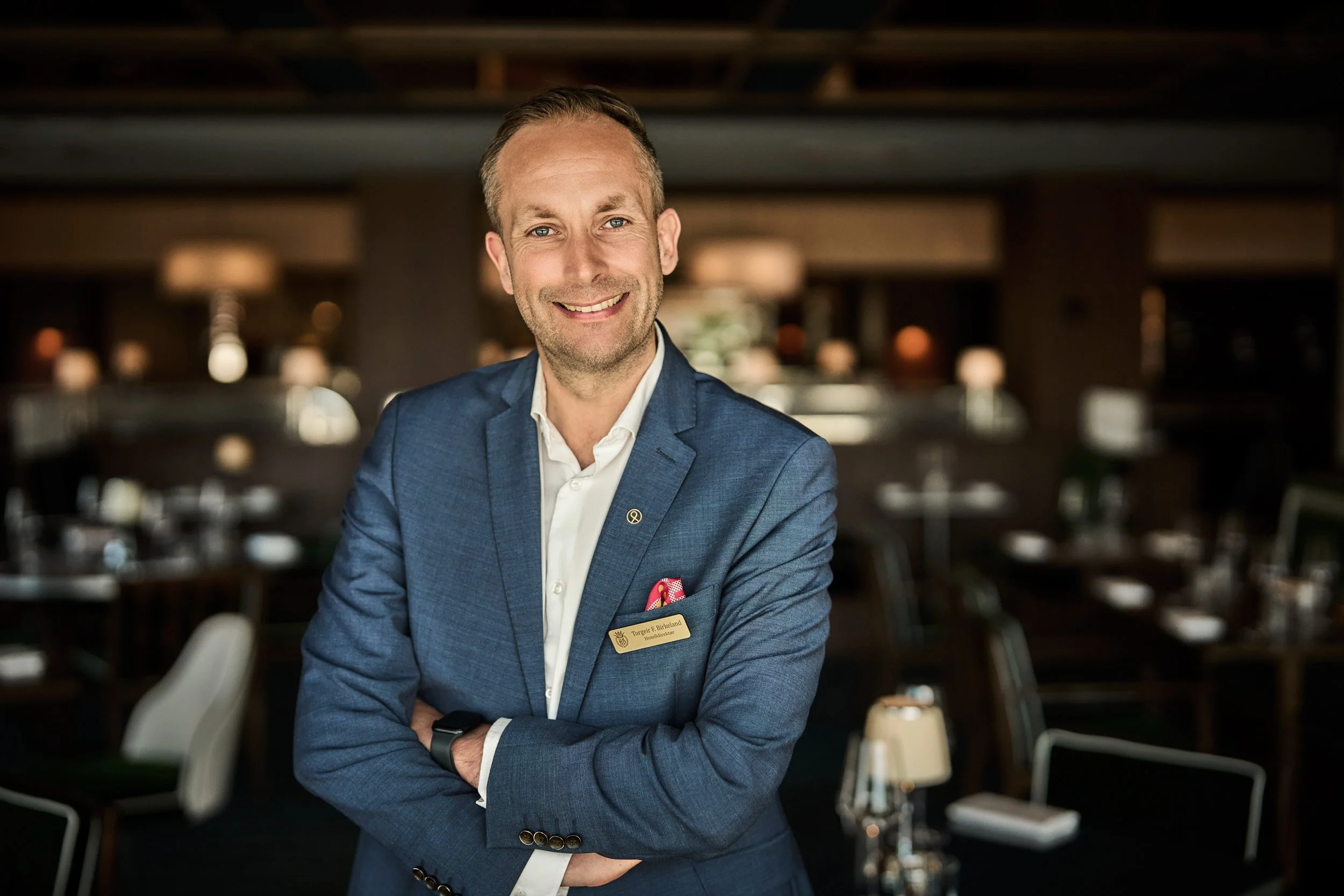 A smiling man in a blue suit with a name tag standing in a restaurant with tables and chairs in the background.