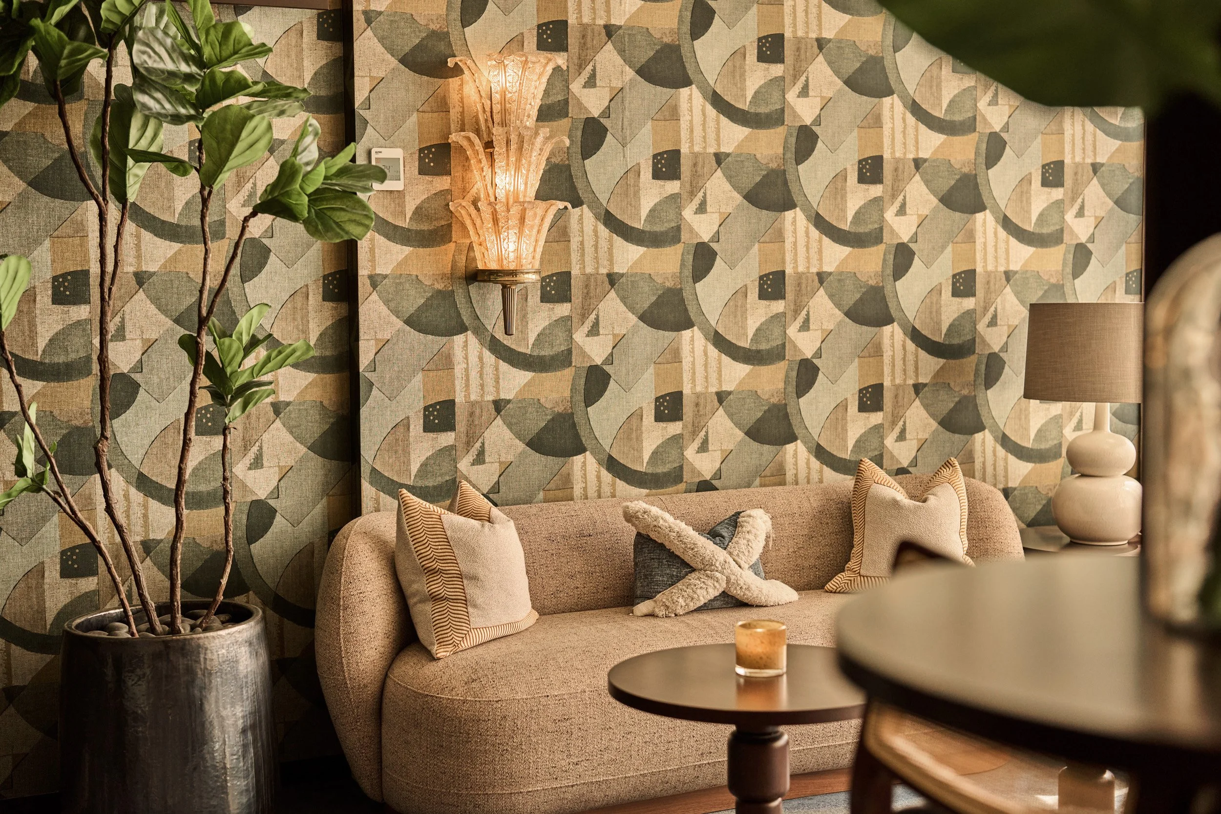 A cozy living room corner with a beige sofa decorated with patterned throw pillows, a dark wooden coffee table with a candle, a large potted plant with tall branches and green leaves, a retro-style wall sconce with a pinkish-glow, a beige table lamp 