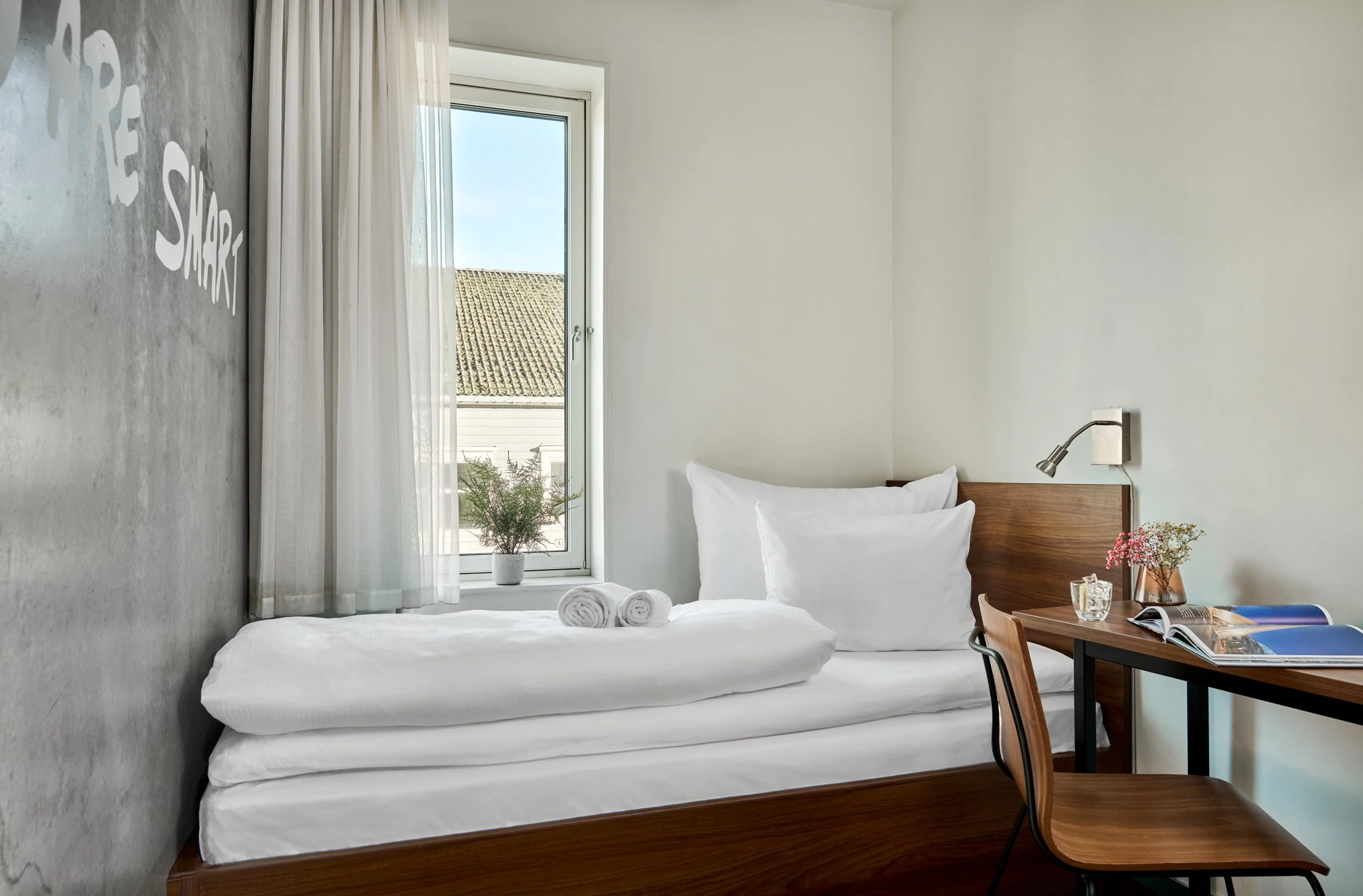 A minimalistic hotel room with a single bed, white linens, pillows, and rolled towels. A window with sheer white curtains shows a view of rooftops and a blue sky. A small desk with books, a glass of water, and a vase of flowers is next to the bed, an
