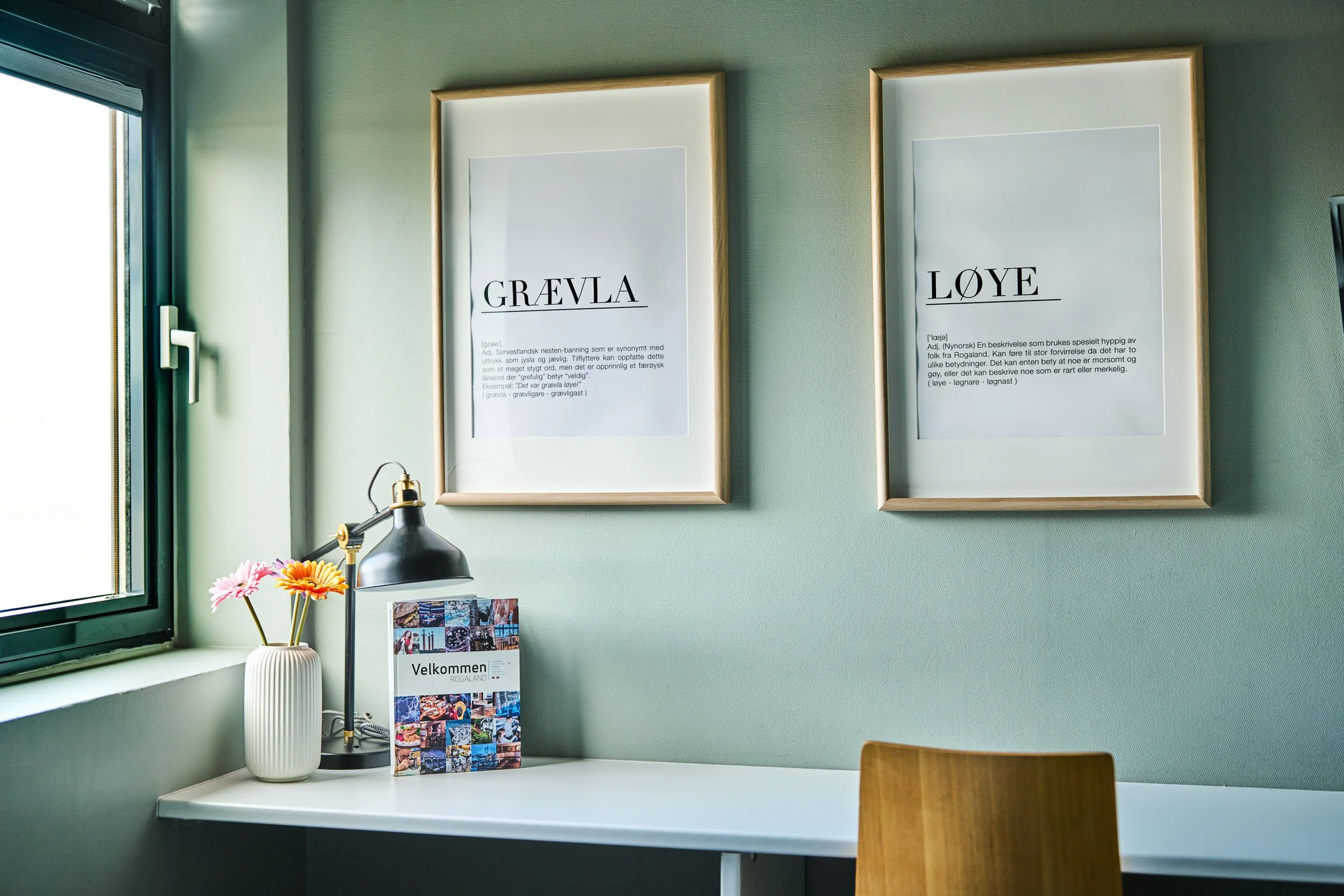A bright room with a window on the left, a white desk, and a wooden chair. On the desk, there is a white vase with pink and orange flowers, a black desk lamp, and a colorful brochure with the word 'Velkommen' on it. The wall behind has two framed pap