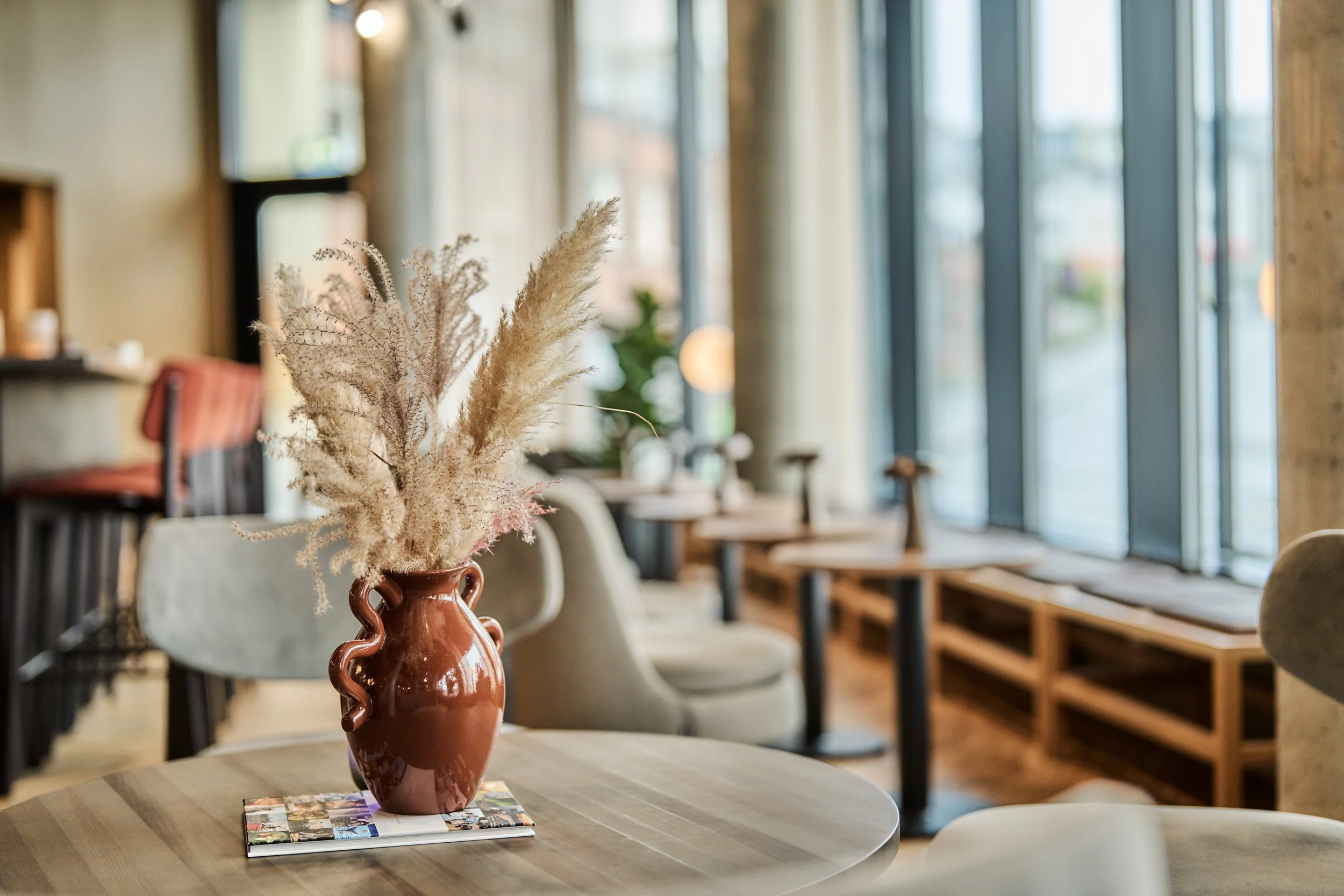 A cozy restaurant interior with a wooden table featuring a brown vase with dried pampas grass, magazines, and soft chairs, illuminated by large windows allowing natural light.