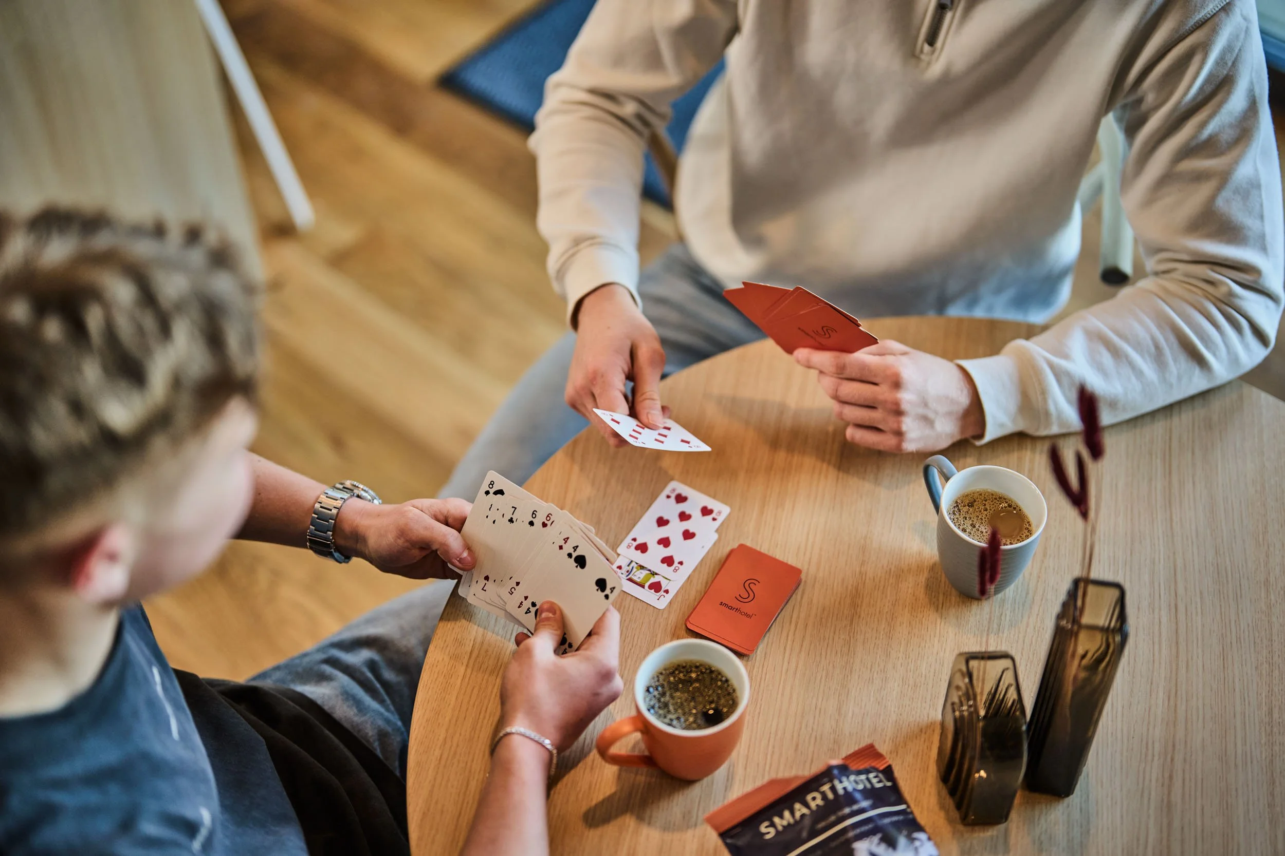 Two people playing cards at a round wooden table with two cups of coffee. Several card cases and a brochure for SMART HOTEL are on the table.