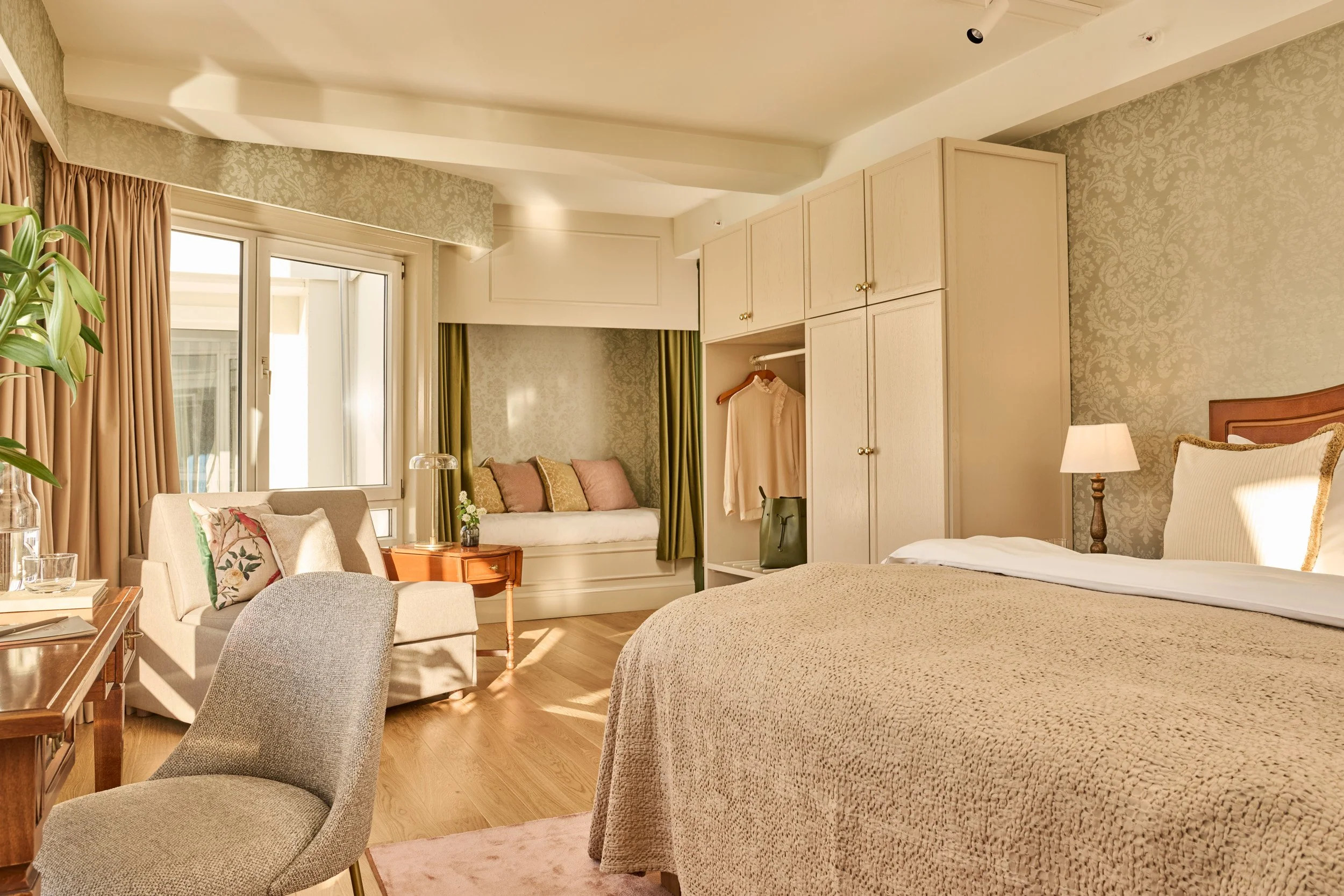 A cozy hotel room at Hotel Ullensvang featuring a bed with beige bedding, a bedside lamp, a built-in window seat with pillows, a beige armchair, a wooden side table, decorated with light-colored wallpaper and curtains.
