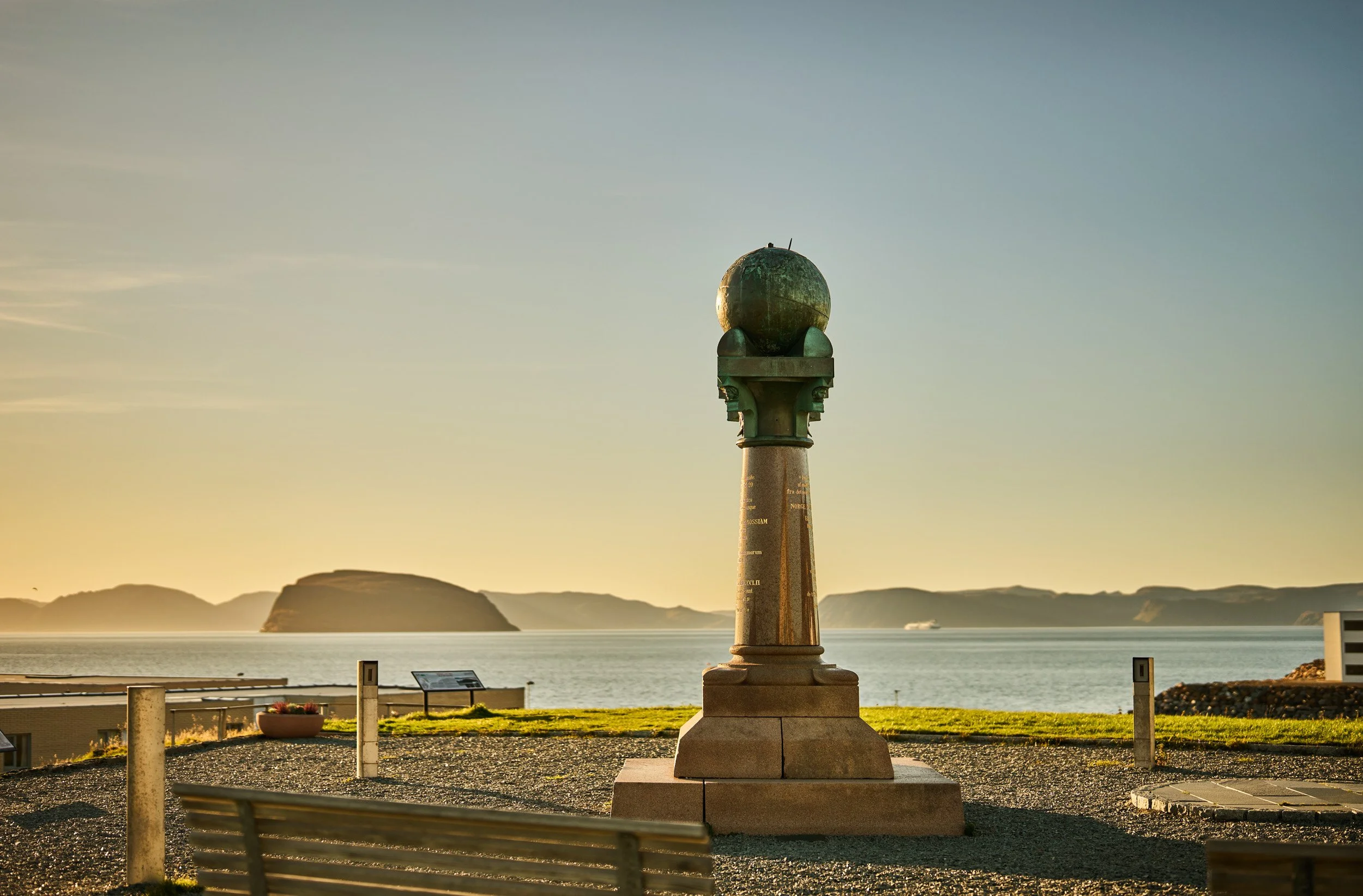 Statue of a globe on a tall pedestal overlooking a body of water at sunset with mountains in the background.