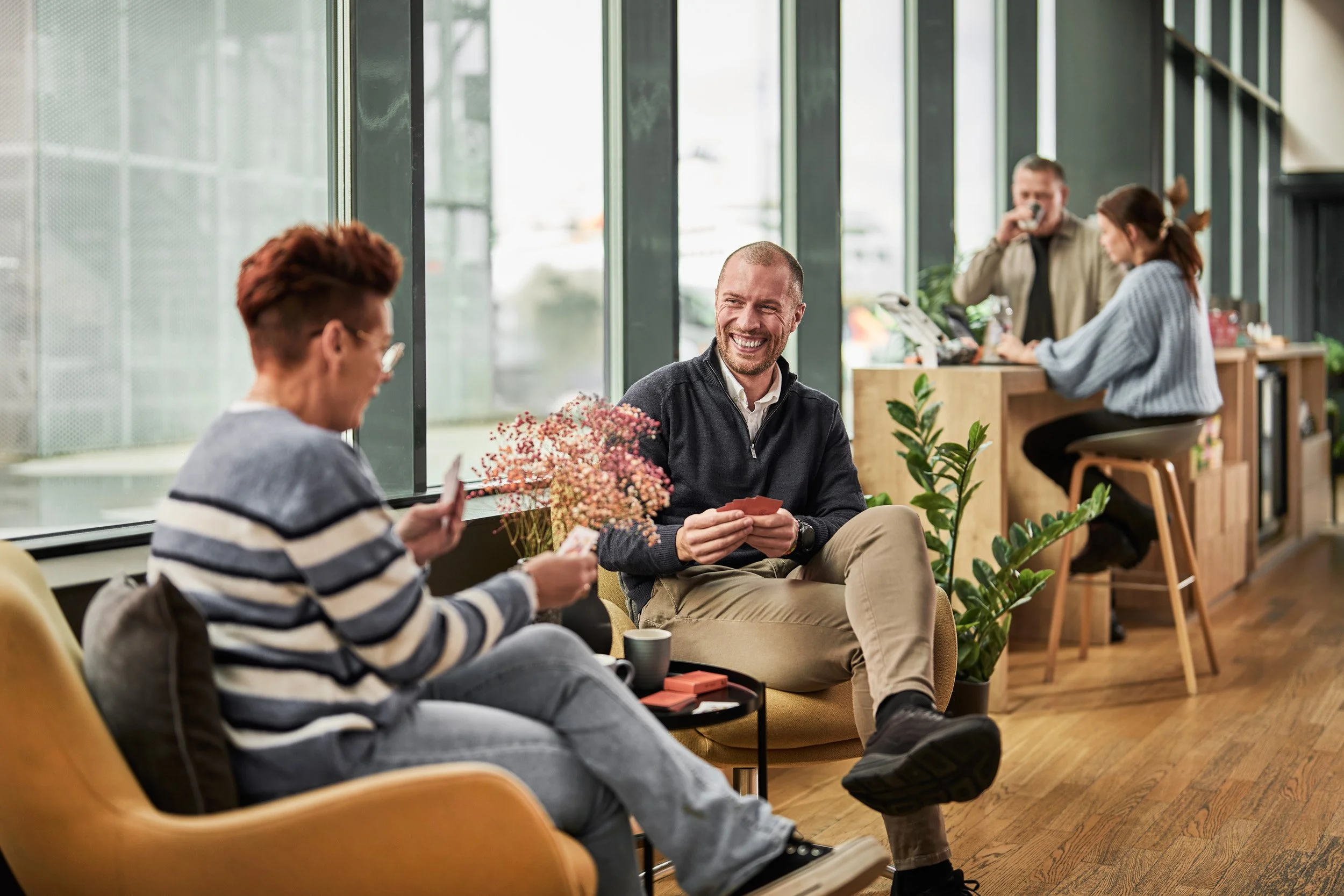 People in a coffee shop, two women playing cards and talking, man laughing, barista preparing drinks in the background, plants and wooden furniture.