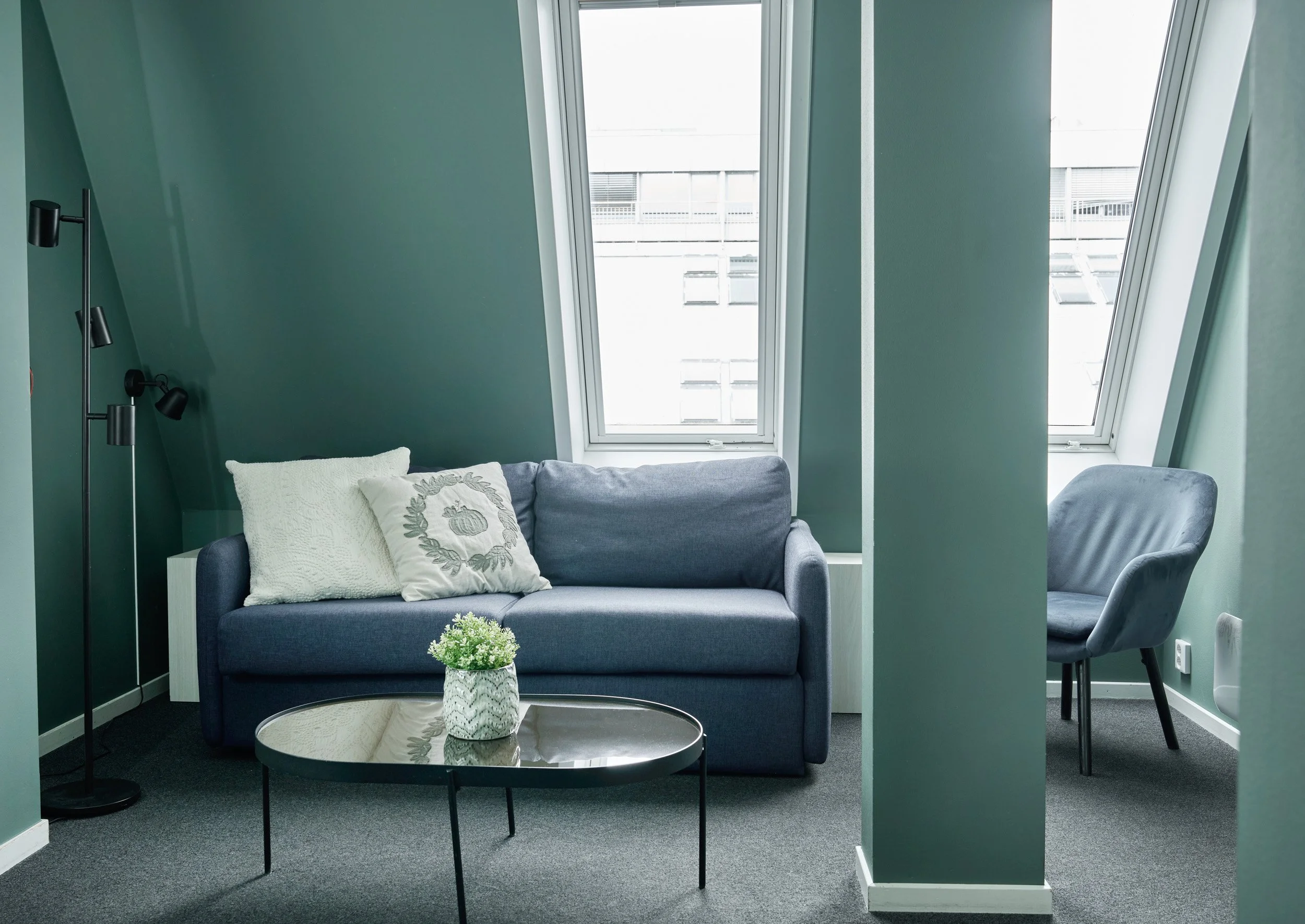 A cozy living room corner with a blue sofa, decorative pillows, a small coffee table with a plant, a black floor lamp, and a gray armchair near two large windows in a green-walled room.