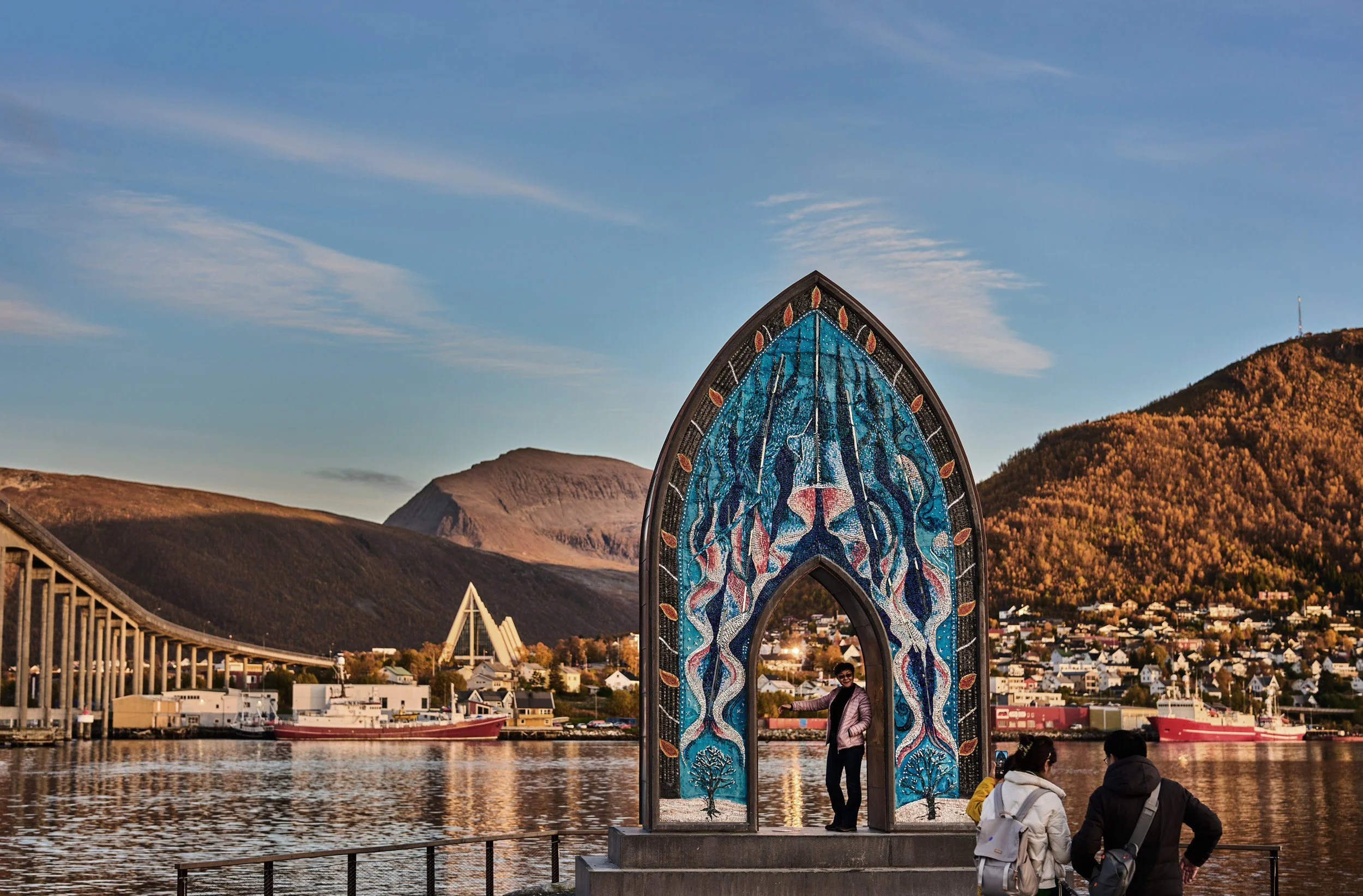 A woman standing inside a colorful stained-glass arch sculpture near the riverbank with mountains and houses in the background, during sunset.
