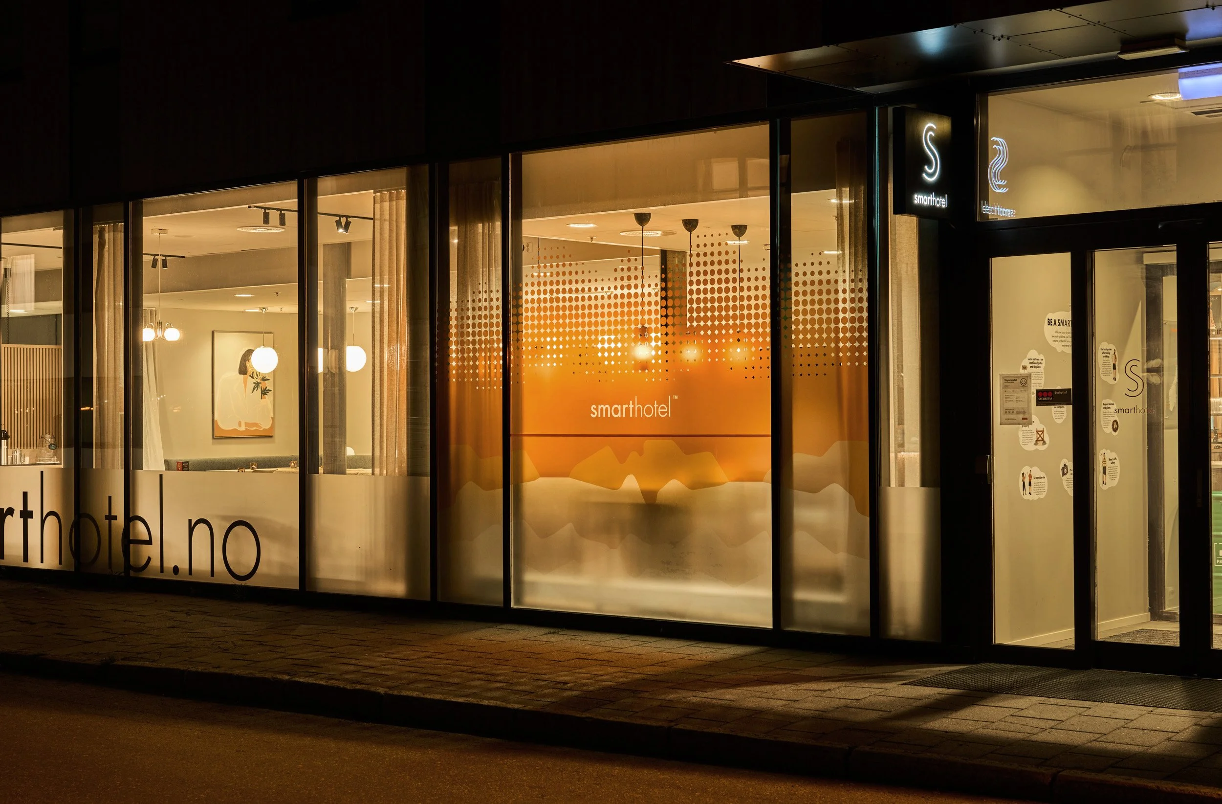 Nighttime exterior view of Smarthotel with illuminated sign, large glass windows revealing a cozy, modern interior with artwork, hanging lights, and curtains.