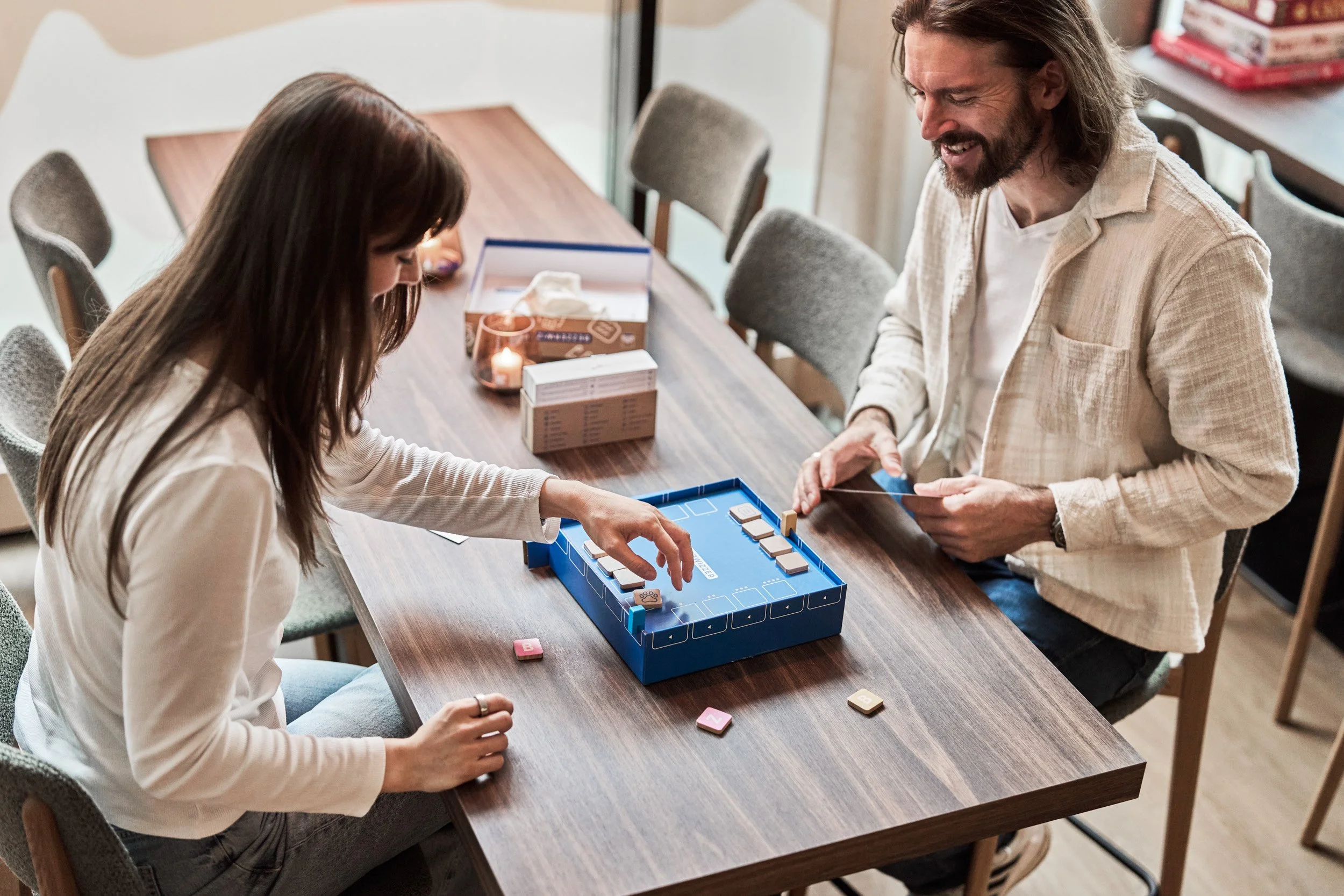 Two people playing a tile-based game at a wooden table, with game pieces and cards, in a cozy indoor setting.