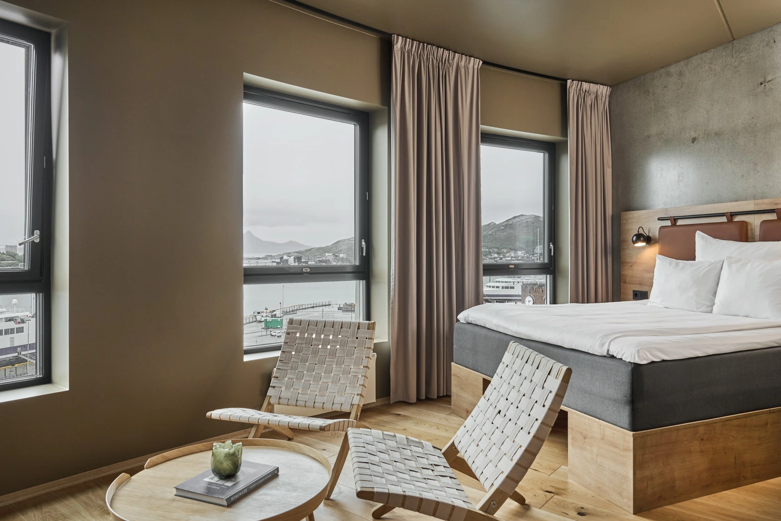 Modern hotel room with large windows, a bed, and two woven chairs around a round table with a green decorative object.